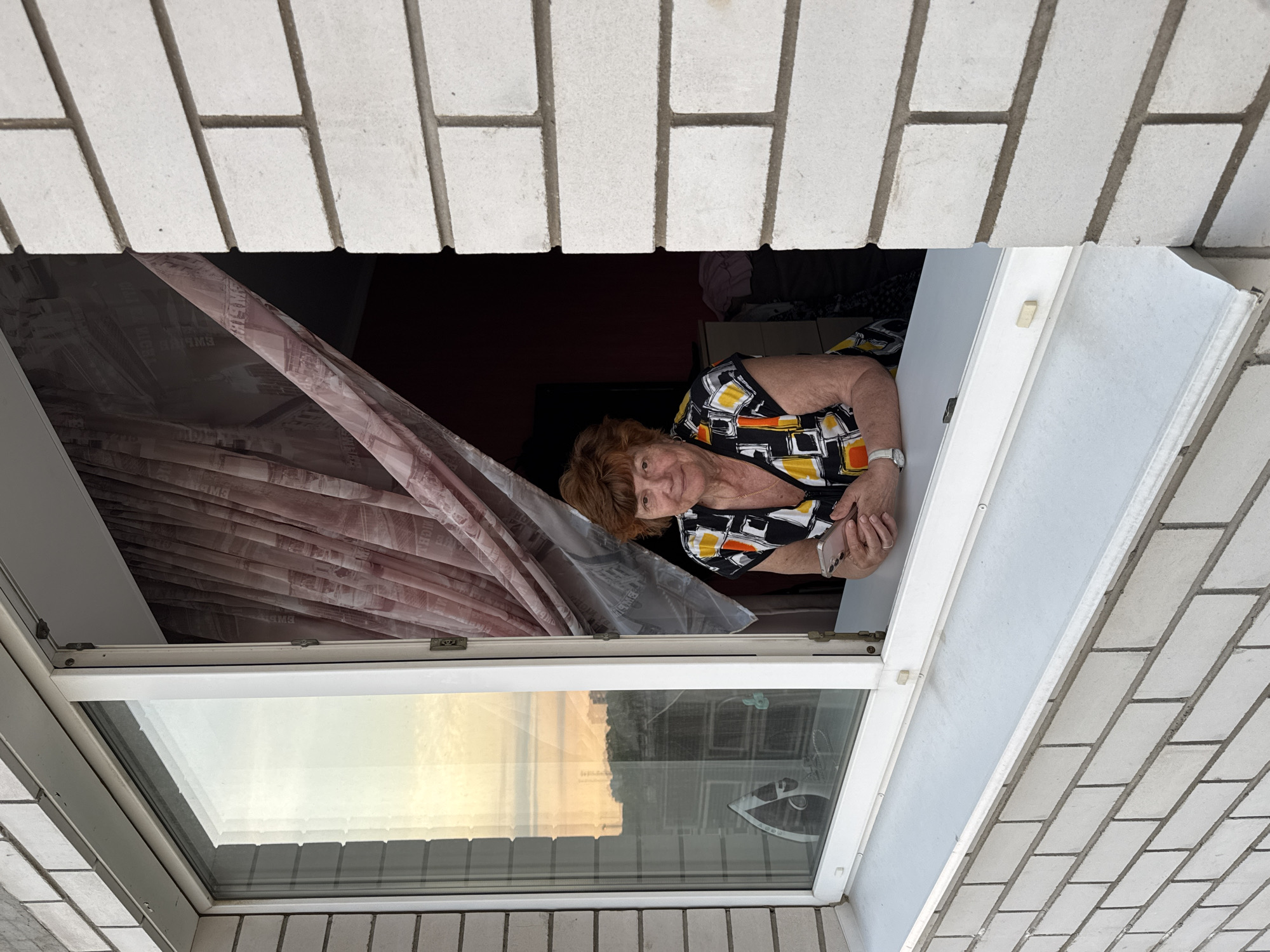 An elderly woman with short, reddish-brown hair and a floral print shirt stands at a partially open window, looking directly at the camera. She is holding a smartphone in her hands, resting her forearms on the windowsill. The woman is positioned in the interior of a room, partially obscured by a sheer, patterned curtain with an image of a city skyline. The window offers a view of the exterior, showing a light-colored brick building surrounding the frame and a hazy sky with hints of orange, suggesting either dawn or dusk. Inside the room, a dresser or cabinet with folded clothes is visible behind the woman. The scene appears to be set on a balcony or in an apartment in Kirishi, Russia, given the context provided.