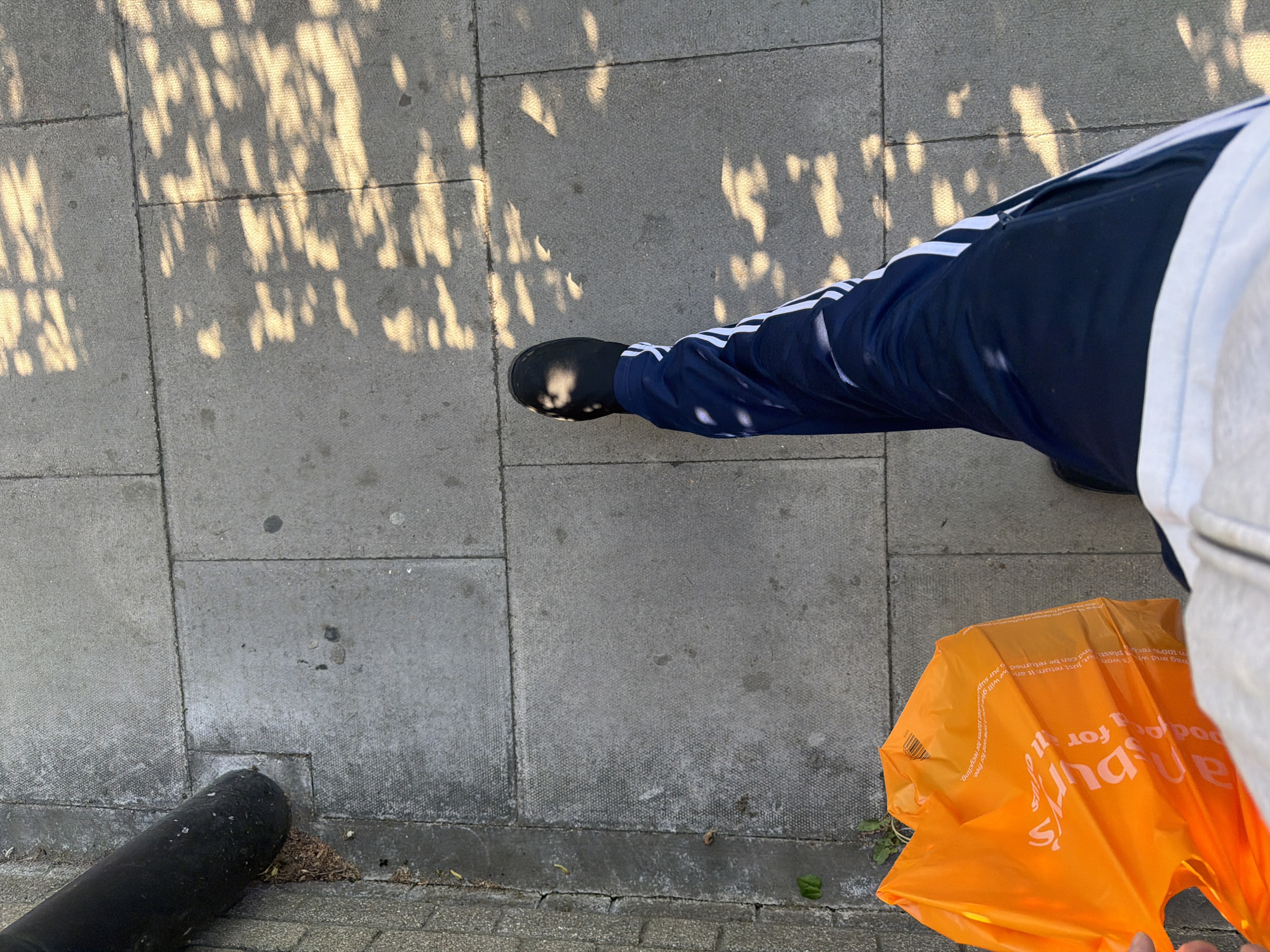 An overhead shot captures a person's lower body standing on a grey paved sidewalk, typical of an urban environment in London, United Kingdom. The ground is composed of rectangular concrete slabs, displaying dappled patterns of sunlight and shadow, suggesting it is a bright, sunny day, possibly morning or late afternoon.
The person is wearing dark blue track pants with distinctive white stripes running down the side, characteristic of Adidas athletic wear. On their right foot, a black, slip-on shoe, resembling a Croc, is visible, with a small patch of sunlight highlighting its surface. To the bottom right, an bright orange plastic shopping bag is held by the person, its crumpled surface revealing partially obscured white text. The text, when read, indicates it's a Sainsbury's bag (a UK supermarket chain), with phrases like "bag and we'll... return it to any... stores for recycling" and "100% recycled plastic" being decipherable.
In the bottom left corner, a black, cylindrical object, possibly a bollard or part of a lamppost, rests on the edge of the pavement. The overall scene depicts a mundane, everyday moment of someone walking or paused on a city street.