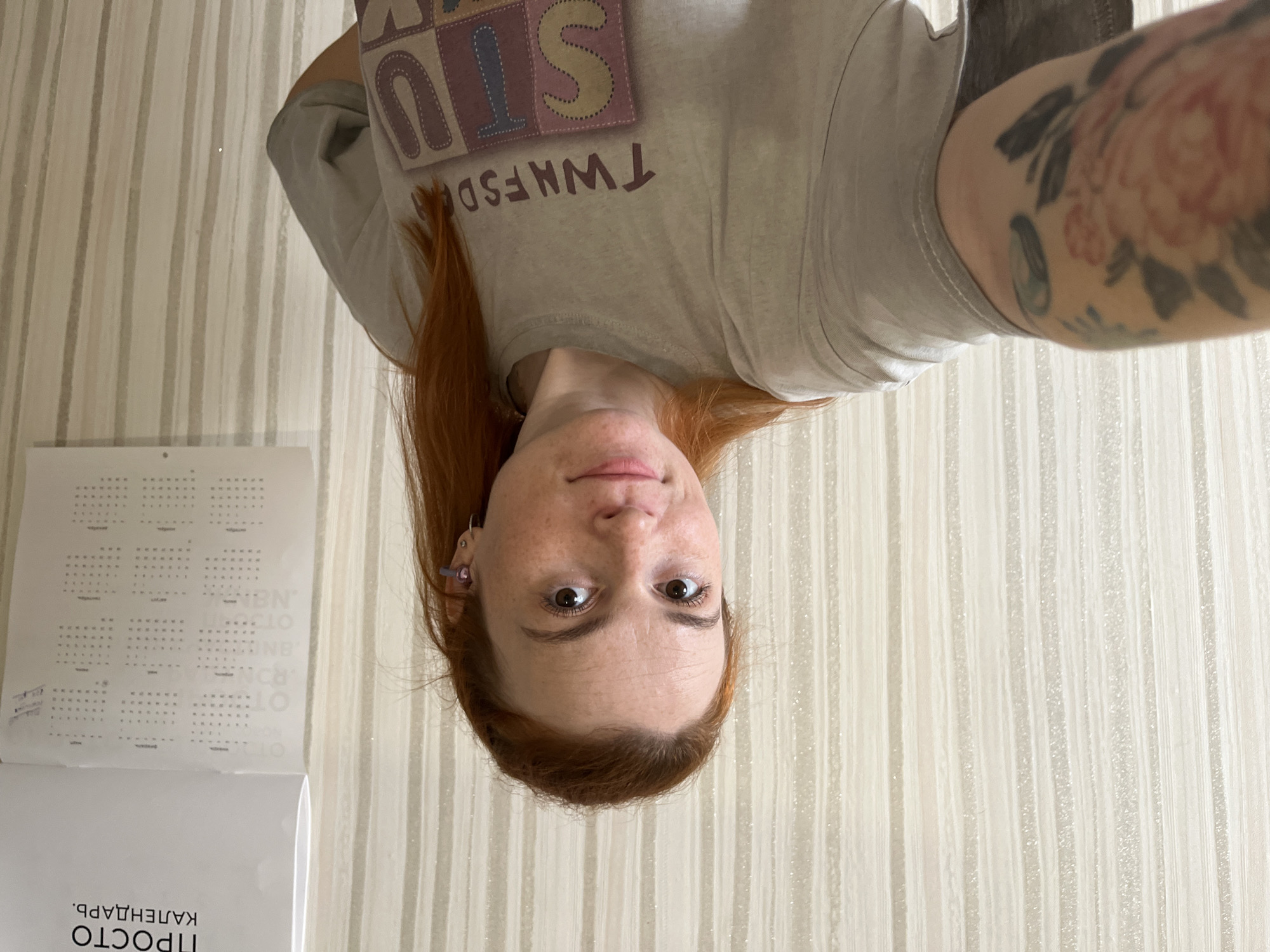 A red-haired woman with freckles and brown eyes takes a selfie in front of a striped beige wallpaper. She is wearing a light grey t-shirt with the text "TWNFSDAY" and a colorful block design. A tattoo of a peony and other designs are visible on her left arm. She has a small purple earring in her right ear. A calendar with Russian text, including "ПРОСТО КАЛЕНДАРЬ" (Simply Calendar), is hanging on the wall to her right. The scene appears to be indoors, possibly in a residential setting in Chaikovsky, Russia.