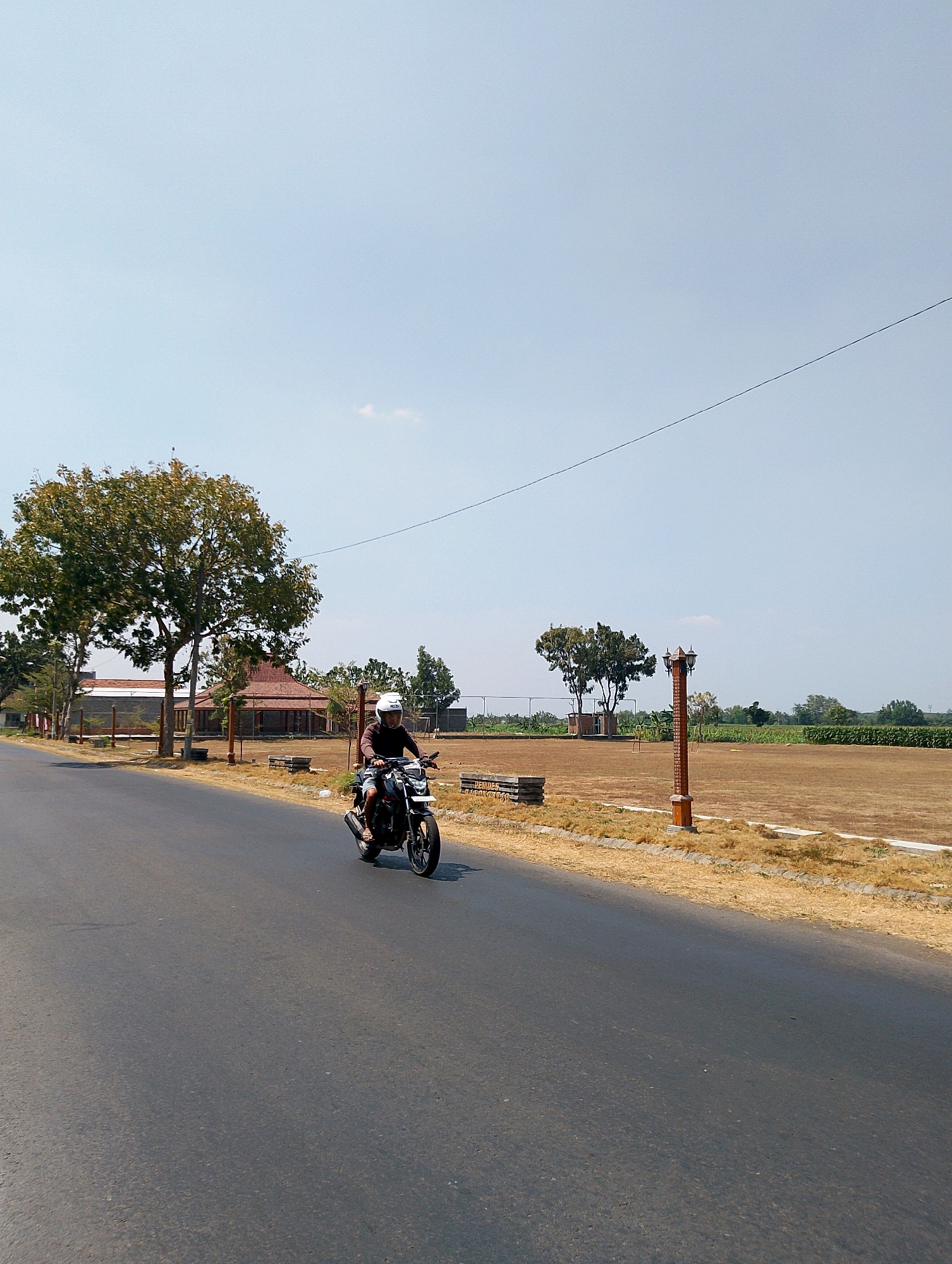 A person wearing a helmet is riding a black motorcycle on a paved road in Balongrejo, Indonesia. The rider is wearing a dark long-sleeved shirt and shorts. The motorcycle has a silver exhaust pipe and black rims. To the left of the road, there are several large trees with green and brown foliage, and a building with a reddish-brown tiled roof. In the background, there is a dry, tan-colored field with some wooden structures and a lamppost. The sky is clear and light blue with a thin wire stretching across it. The overall environment suggests a rural or semi-rural area.