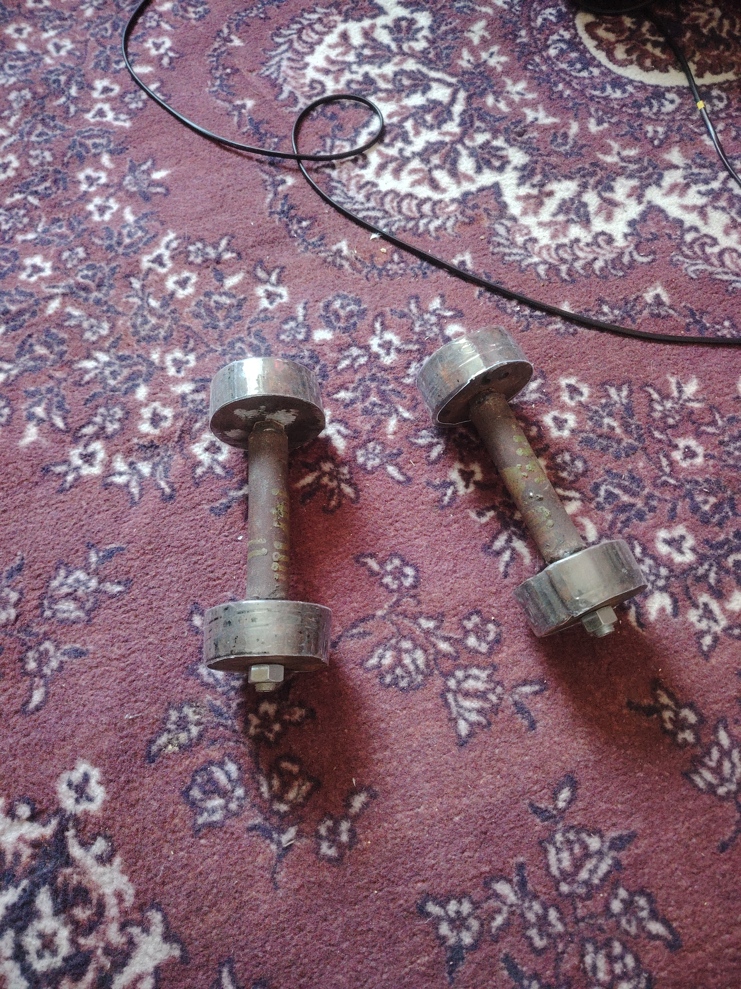 Two dumbbells are lying on a maroon carpet with a floral pattern. A black cable is visible in the upper right portion of the image, partially looped. The dumbbells are metallic and appear to be old or well-used, with some rust or discoloration on the handles. The weights at the ends of the dumbbells are cylindrical and have a polished finish. The scene is indoors, as indicated by the carpet and the presence of the cable. The location is specified as Александров ауылдық округі, Kazakhstan.