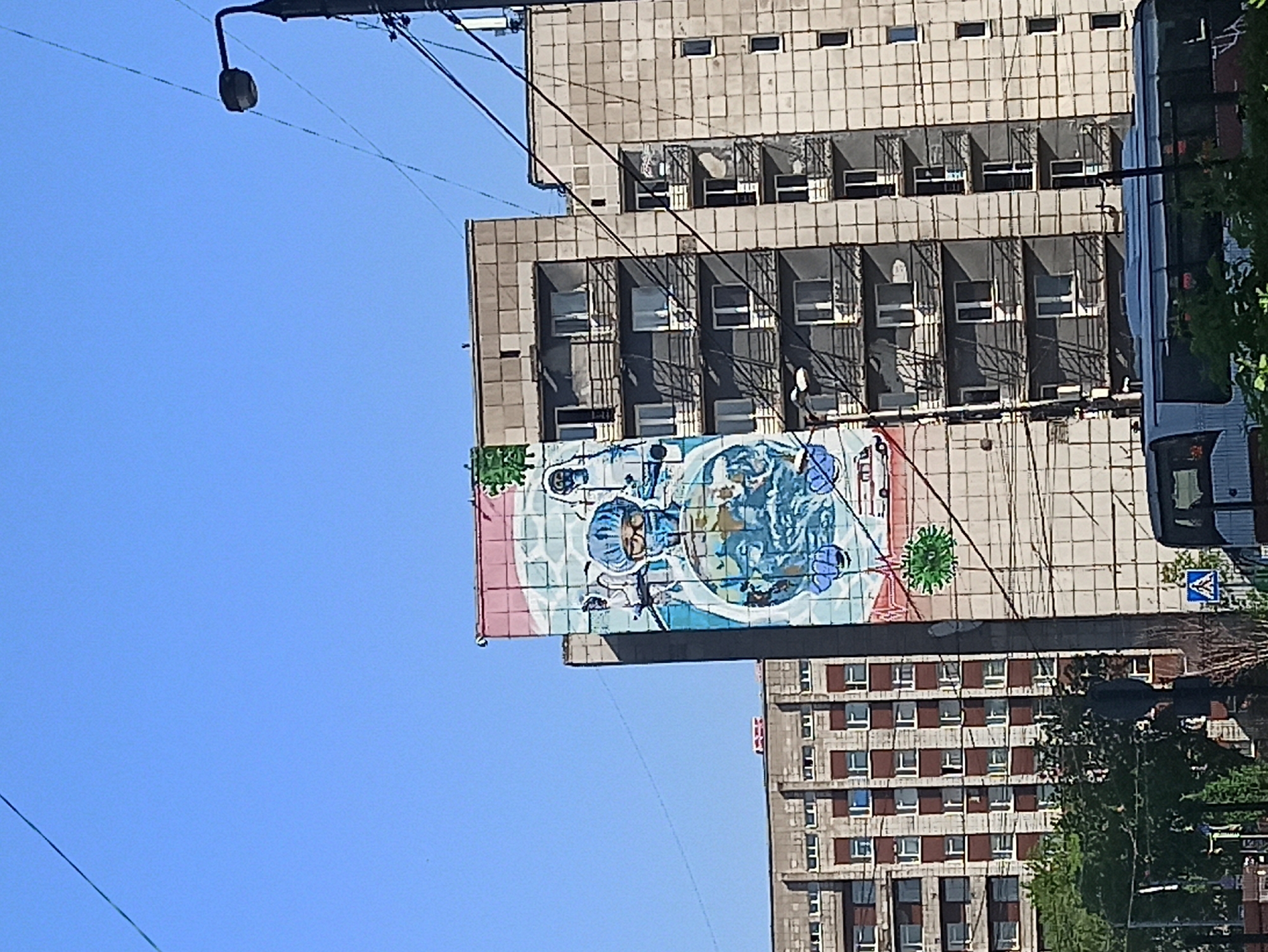 The image shows a building in an urban area. The building is made of grey concrete tiles and has several windows. A large mural is painted on the side of the building, depicting an animated figure with blue hair and glasses, surrounded by other blue and white shapes. A second building can be seen in the background with brown-framed windows. Green trees and a bus are visible on the right side of the image. Overhead power lines and a street light are also present. The sky is clear and blue. The location is Unknown, Unknown.