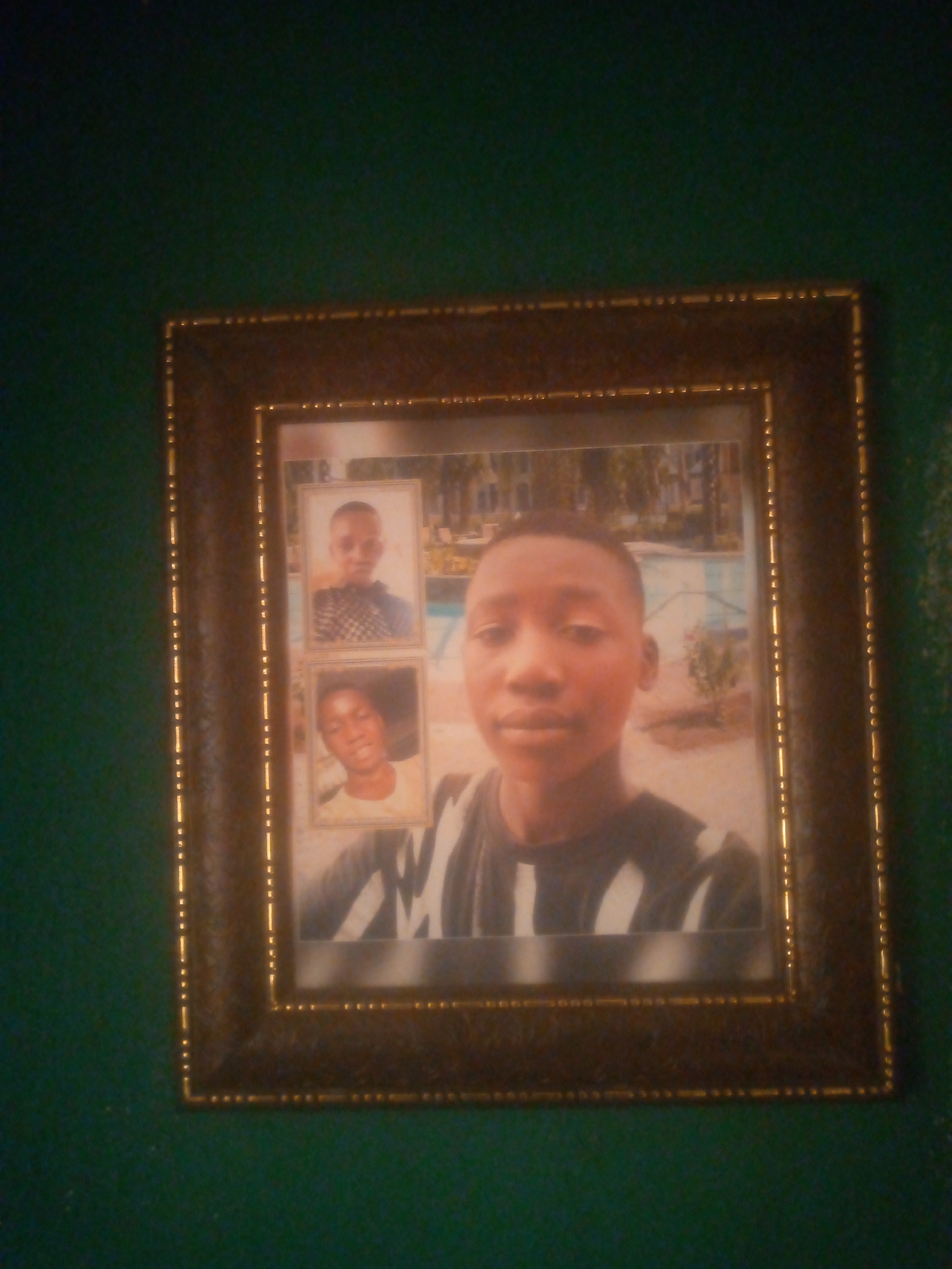 A framed photograph hangs on a green wall. The photograph contains three separate images of a young, dark-skinned male. The main image features him wearing a black and white striped shirt, with a background that suggests an outdoor location with a swimming pool and vegetation. Above and to the left of the main image are two smaller, separate portraits of the same individual, one with a darker patterned shirt and another with a light yellow shirt. The location is described as Unknown, Unknown.