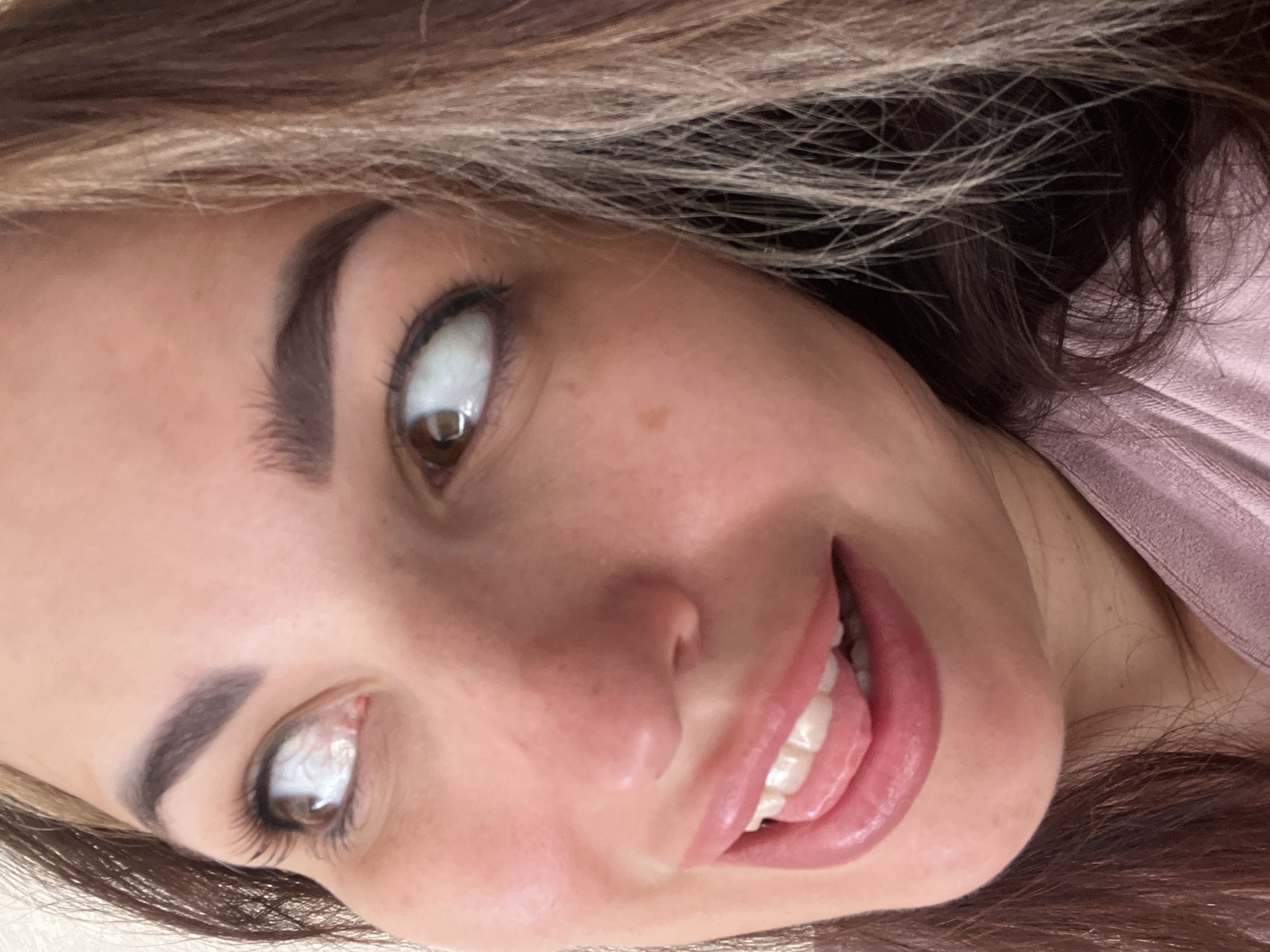 A close-up, eye-level, over-the-shoulder shot captures the face of a woman, cropped at the neck and partially obscured by her hair and clothing. Her head is tilted upwards and to her right, with her eyes also looking upwards and to her right. She has brown hair with blonde highlights, dark eyebrows, and is wearing a pink top. Her mouth is open, with her tongue sticking out and touching her lower lip. The background is a plain, light-colored wall. The lighting is soft and natural.