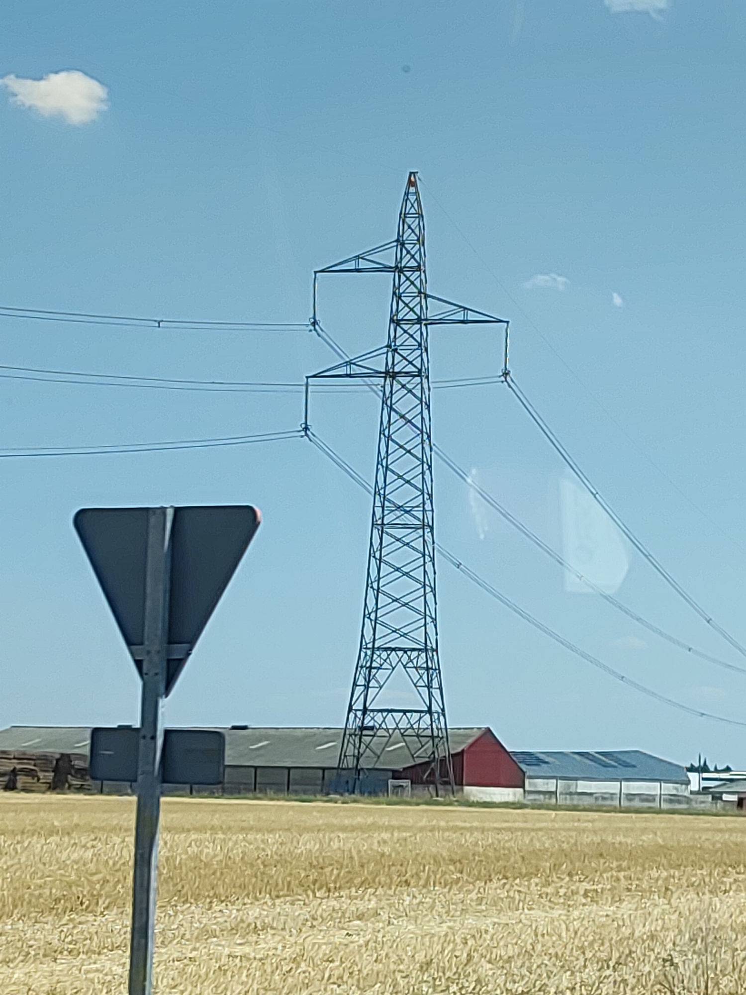 The image shows a rural landscape under a clear blue sky with a few small clouds. Dominating the center is a large, lattice-style electrical pylon with multiple power lines stretching across the frame. In the foreground, to the left, is a grey, triangular road sign mounted on a metal pole. Behind the sign and the pylon, a field of dry, golden crops, likely wheat or barley, stretches into the distance. A row of agricultural buildings with dark, sloped roofs is visible behind the field. One building is painted red, and others appear to be long, low structures with white walls and multiple openings. The scene is consistent with an agricultural area in Cogeces del Monte, Spain. There are no people visible in the image.