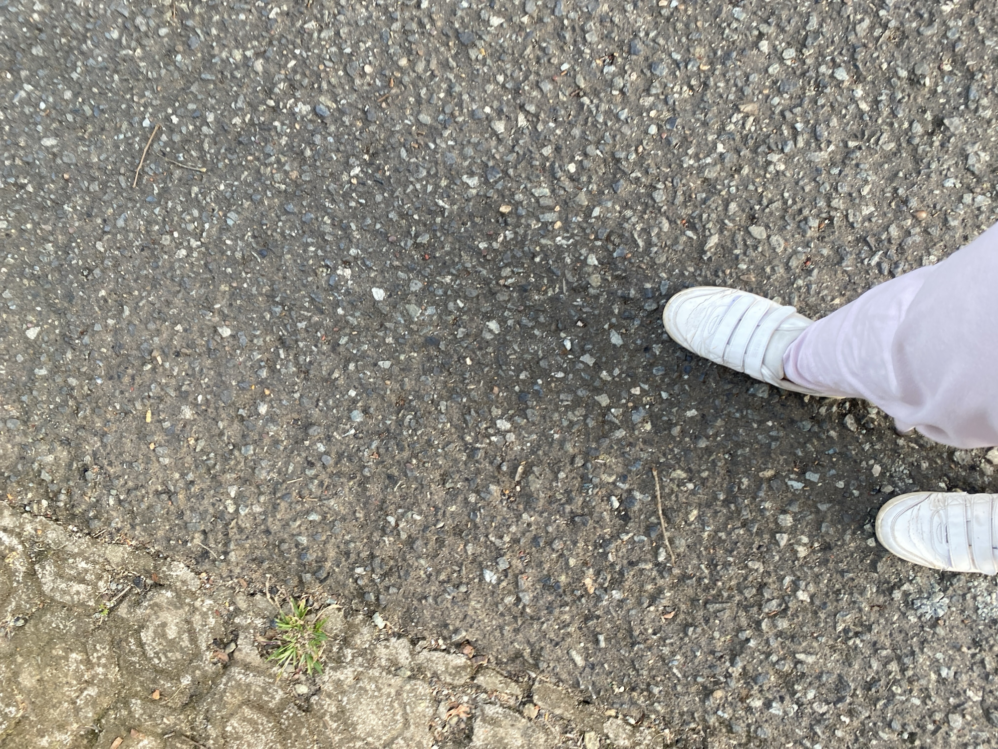 A first-person perspective shot shows the feet of a person wearing white sneakers and light pink pants. The person is standing on a dark asphalt surface with small grey and white pebbles embedded in it. To the left, the asphalt meets a cracked concrete surface, also with pebbles. A small patch of green grass is visible growing from a crack in the concrete. The scene appears to be outdoors, possibly on a street or pathway. The lighting suggests it is daytime. The location is identified as Banjarbaru, Indonesia.