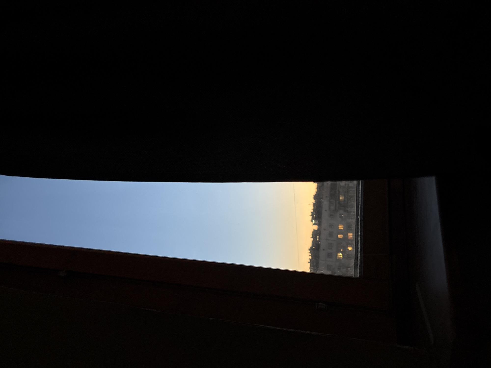 The image captures a view through a window, possibly a skylight or attic window, from a darkened interior space. The window is framed by dark, indistinct material above and to the sides, with a wooden sill or frame along the bottom.
Through the window, a clear sky is visible, transitioning in color from a light blue on the left to a warm, soft yellowish-orange on the right. This gradient suggests either a sunrise or sunset.
On the far right side, partially obscured by the window frame and the darkening sky, a distant, multi-story building complex is visible. The architectural style, with its grid of windows, appears characteristic of residential or apartment blocks often found in urban or suburban areas, consistent with the provided location context of Russia. Several windows in this distant building are illuminated with warm, yellow lights, further emphasizing the idea of evening or early morning. There are also subtle reflections of these lights within the windowpane.
The immediate surroundings within the room are dark and out of focus, drawing attention to the framed view outside. The scene conveys a sense of quiet observation from an indoor vantage point, capturing a transitional time of day. No people or visible text are present in the image.