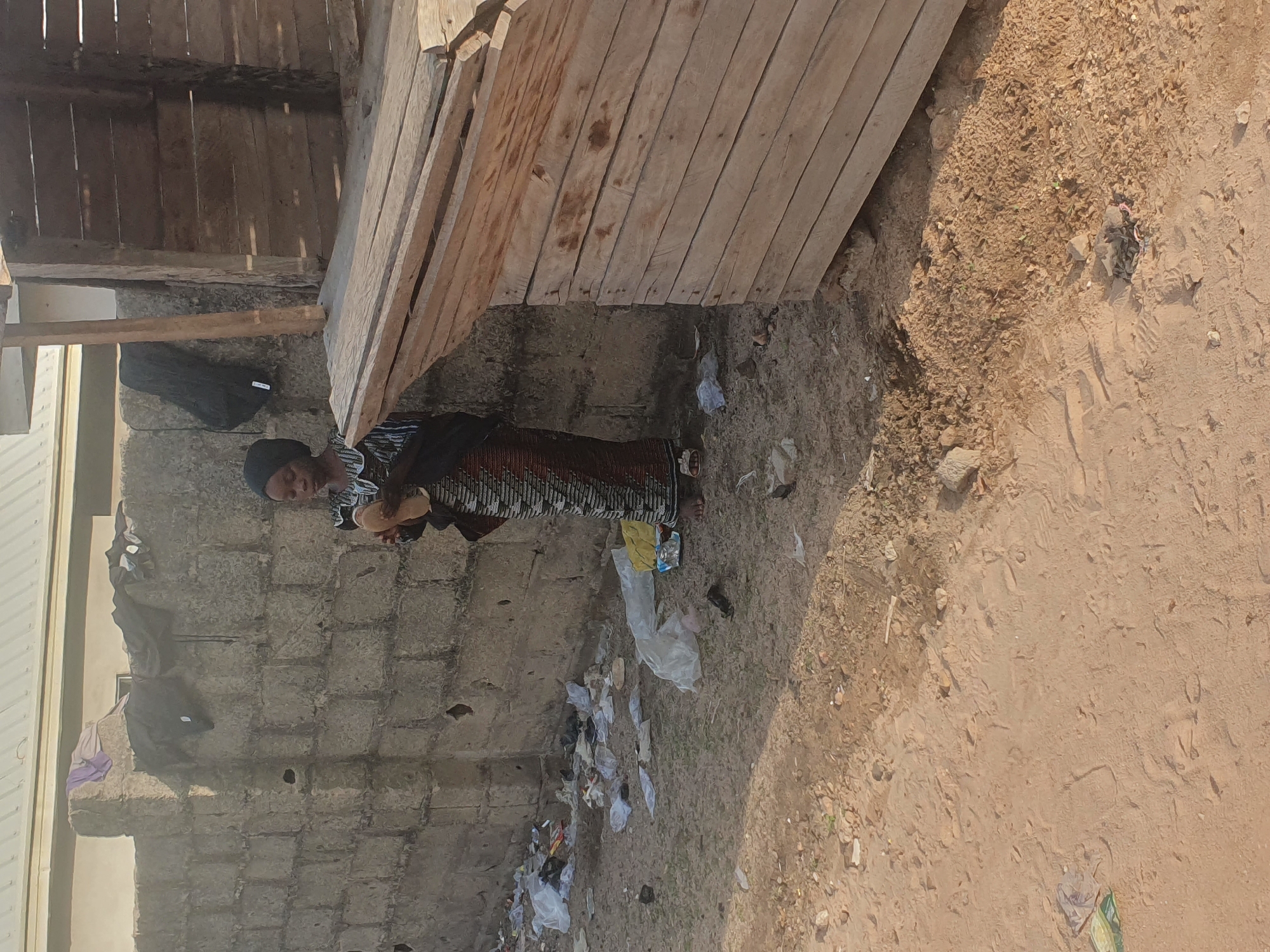 The image depicts an outdoor scene in Lokoja, Nigeria. An individual stands on a patch of dirt and debris next to a cinderblock wall. The person is wearing a dark head covering, a patterned top, and a long skirt or dress. They appear to be holding a light-colored object in their hands. Above the wall, there are wooden planks, partially obscuring the scene. The ground is a mixture of dirt and scattered trash. The scene appears to be in a somewhat impoverished or neglected area.