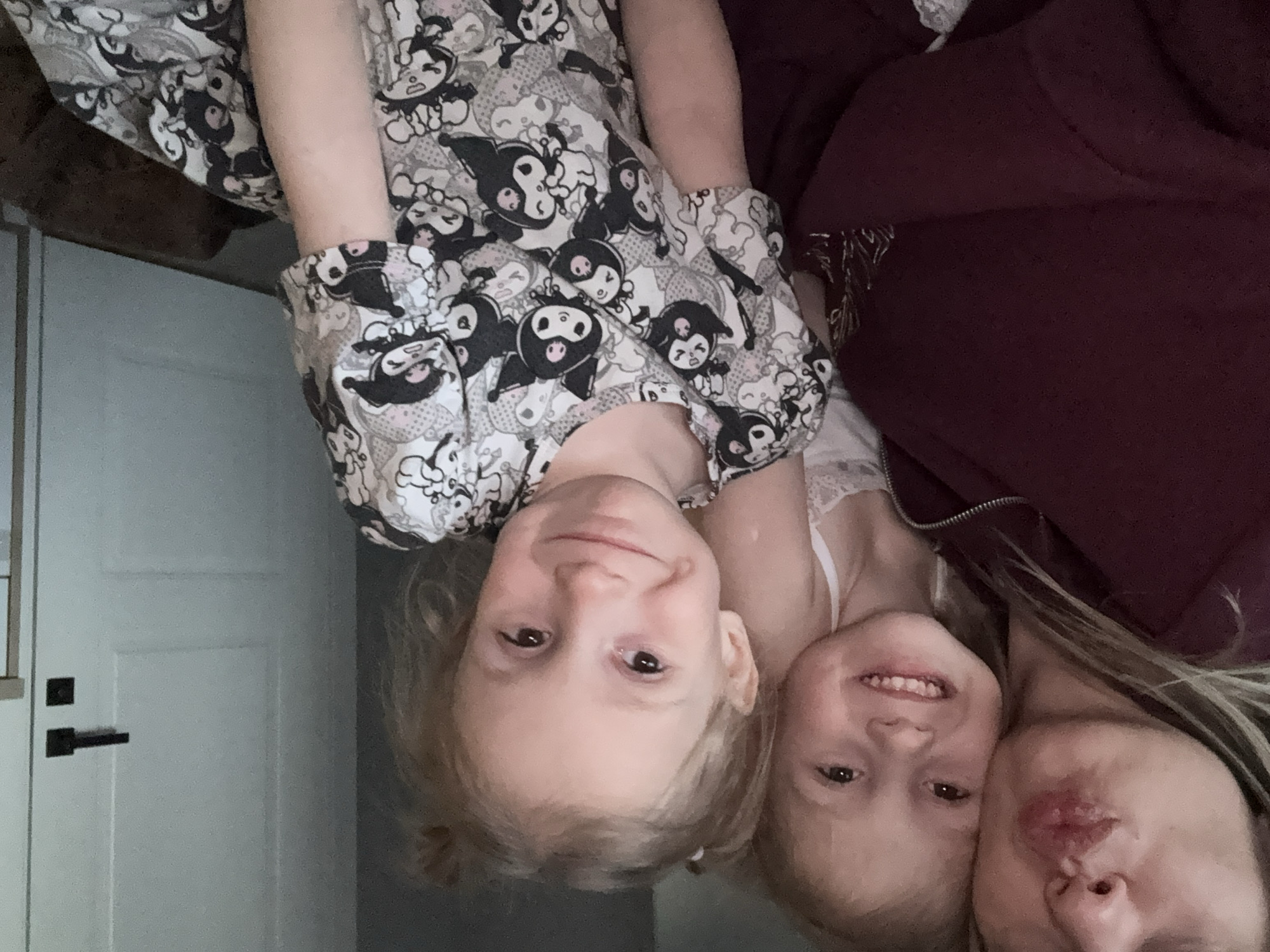 The image depicts three individuals in an indoor setting. A young child, wearing a patterned grey and white top with dark cartoon characters, is positioned at the top left. Another child, wearing a white top with lace trim, is located in the middle. Adjacent to her is an adult with long blond hair wearing a dark burgundy or maroon colored hoodie. The background includes a white door with a black handle and door hardware. The location cannot be identified as Krasnoyarsk, Russia.