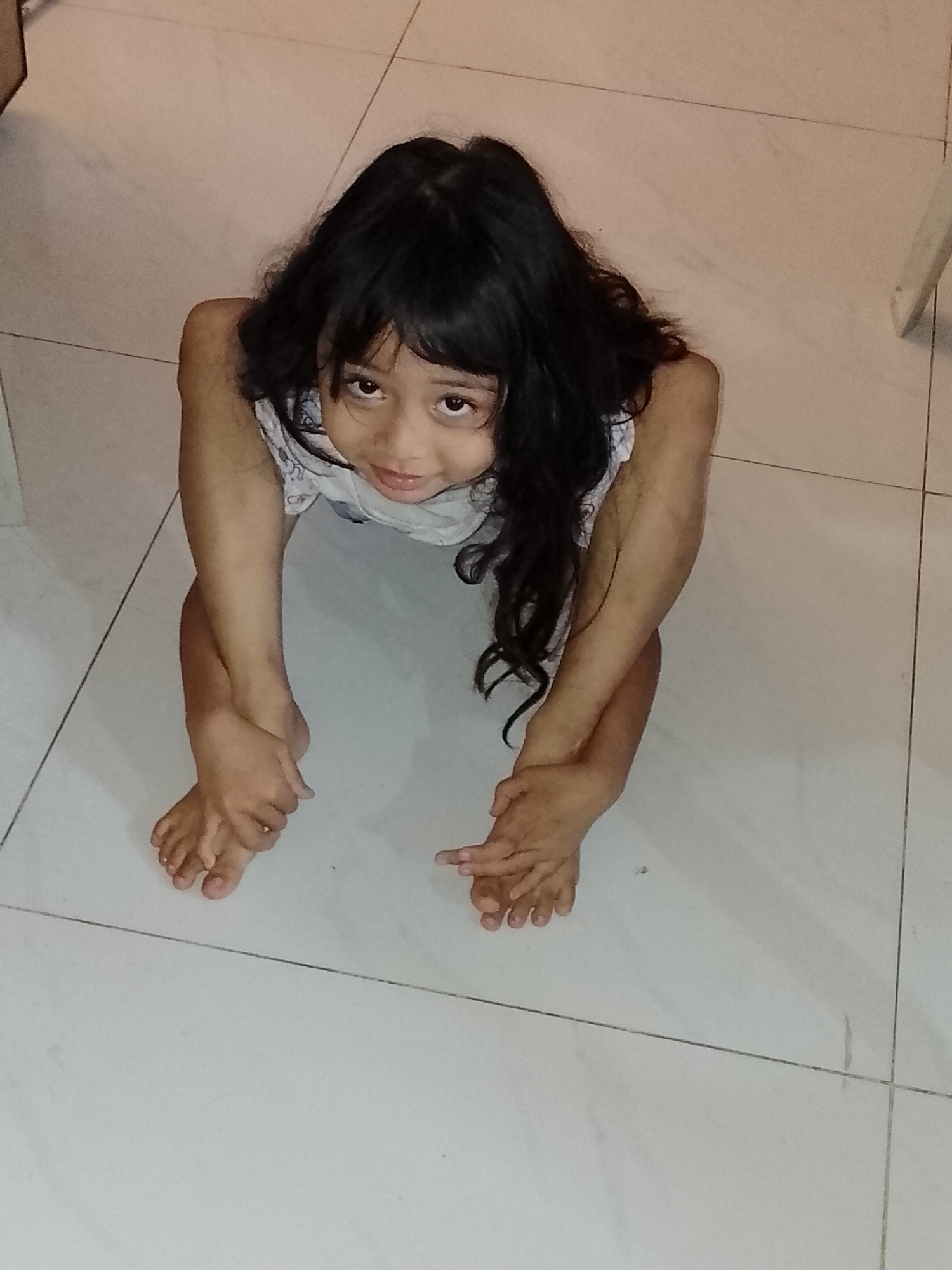 A young girl with long, dark, curly hair and brown eyes is centered in the frame, looking directly up at the camera with a gentle, curious expression and a slight smile. She has a medium-brown skin tone and is wearing a light-colored, possibly patterned, sleeveless top or dress.
The girl is in a crouching or squatting position on a light-colored tiled floor, with her legs bent and drawn close to her body. Her arms are bent, and her hands are positioned near her feet. One hand appears to be holding or resting on her foot. The tiles are large and square, with dark grout lines that run horizontally and vertically across the floor, creating a grid pattern.
The setting is indoors, with uniform, bright lighting that suggests daytime. Hints of other structures, possibly furniture or parts of a wall, are visible in the top left and right edges of the image. The overall impression is an intimate, engaging portrait of a child in a domestic setting within Davao City, Philippines. There is no visible text in the image.
