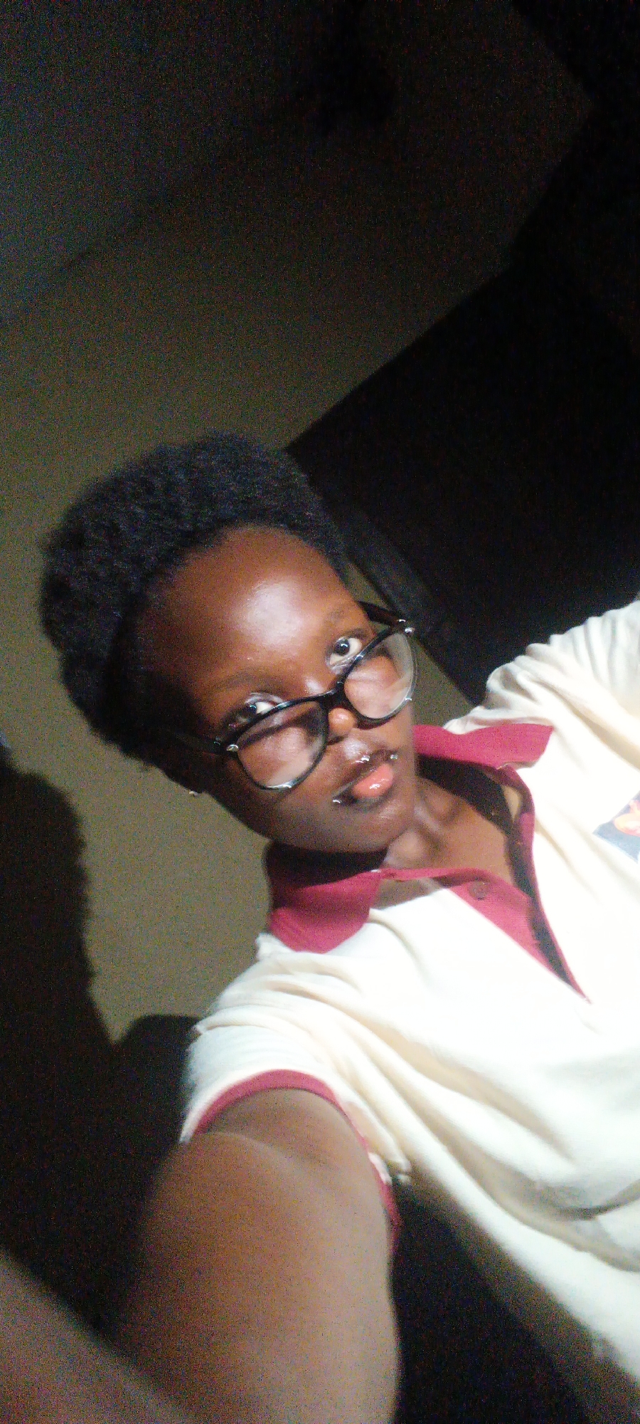 A dark-skinned person with short black hair and glasses is taking a selfie. They have lip piercings and are wearing a light-colored polo shirt with a red collar. The background is dimly lit, with a dark area behind and to the right of the person and a lighter-colored wall to the left. The person is positioned at an angle to the camera. The lighting is uneven, with the left side of the person's face appearing brighter than the right. The environment appears to be indoors. There is no specific information to identify a location in Onitsha, Nigeria.