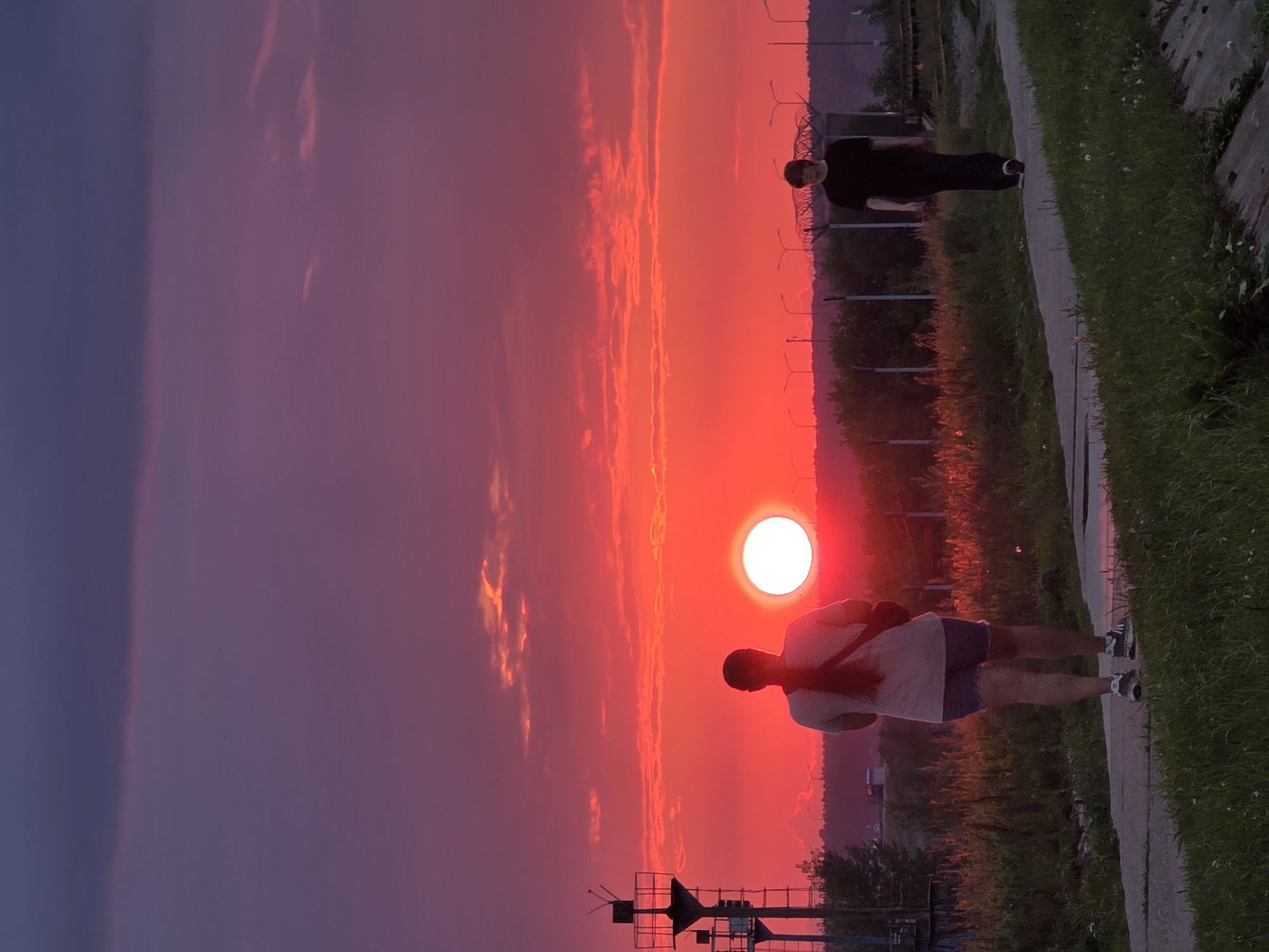 An image captures a vivid sunset over Chaikovsky, Russia, painting the sky with intense shades of red and purple. In the foreground, two individuals are walking along a paved path. A person with long, dark hair, wearing a light-colored t-shirt and denim shorts, is walking away from the camera. To their right and further down the path, a person dressed in black is walking towards the camera. The sun, a bright white orb, is prominently visible near the center of the frame, casting a fiery glow across the horizon. A tall industrial structure, resembling a lighthouse or a communication tower, stands to the left of the image. The background features a line of trees and distant streetlights. The scene is set amidst a natural landscape with overgrown grass and vegetation lining the path.