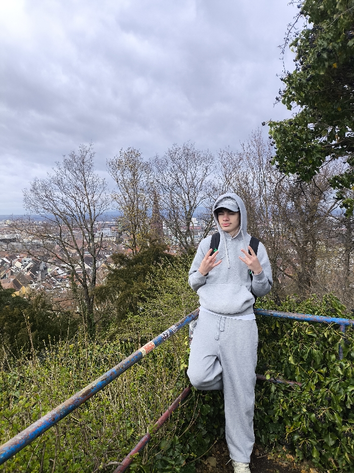 An outdoor, full-length image depicts a young male posing on an elevated viewpoint, likely on the Schloßberg, overlooking the city of Freiburg im Breisgau, Germany. The individual, appearing to be in his late teens or early twenties, is dressed in a matching grey hooded tracksuit; the hood is up, covering a grey patterned bandana or cap. He wears a dark-colored backpack with distinct green straps over his grey hoodie, and a white undershirt is visible beneath the hoodie's hem. His footwear consists of light-colored, possibly beige or off-white, shoes. Both of his hands are raised to chest level, with the index and middle fingers extended in a 'V' gesture. He leans with his right side against a weathered metal railing, painted blue and red, with several stickers affixed to its surface, which runs diagonally through the lower portion of the frame. The foreground and midground are characterized by dense green foliage, including ivy, and numerous deciduous trees, many still bare or with nascent leaves, indicative of late winter or early spring. Beyond the trees, the cityscape of Freiburg im Breisgau stretches into the background under a uniformly overcast sky, revealing a dense urban fabric of buildings, many with red-tiled roofs, and a prominent, partially obscured cathedral spire.