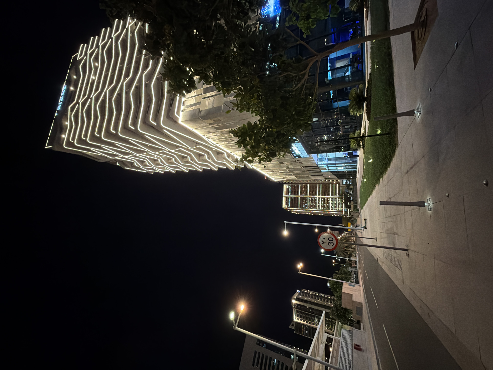 The image captures a modern urban landscape at night in Lusail, Qatar, showcasing illuminated buildings and a quiet street scene.
The dominant subject is a tall, contemporary building on the left, whose facade is spectacularly lit with numerous bright, wavy LED light strips that follow an intricate pattern. On the upper left side of this building, light blue text is visible, appearing to spell "TRIANE" or a similar name. In the mid-ground, a large tree with dark foliage partially obscures the illuminated building.
To the right, a paved sidewalk stretches alongside a dark road, which appears largely empty of traffic. Multiple streetlights with warm yellow and bright white glows line the street, casting light onto the ground and other structures. A prominent red circular traffic sign displays the number "30" in white, indicating a speed limit, with additional Arabic text positioned below it. Further along the sidewalk, sections of green grass and small palm trees are visible, subtly lit by ground-level fixtures. In the background, other high-rise buildings with illuminated windows contribute to the city's nighttime glow.
The scene depicts a clear night. There is no visible human activity. The overall impression is one of modern urban development, tranquility, and striking architectural lighting.