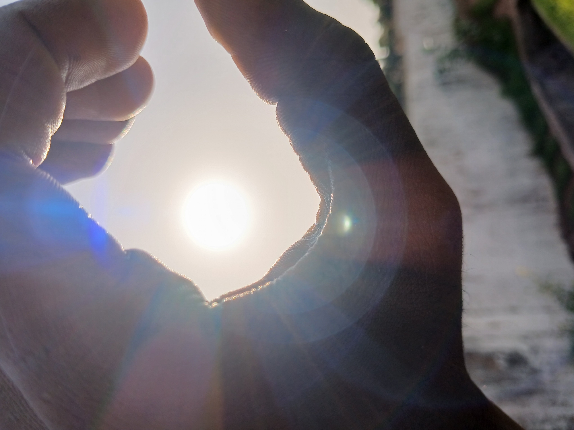 The image is a close-up, low-angle shot of two hands forming a heart shape, with the sun shining brightly through the center. The hands are dark-skinned and appear to be adult. The sun is a brilliant white orb, creating lens flare and light streaks across the image, with distinct blue and purple hues visible. The background is out of focus, showing a blurred light-colored wall and some greenery. The scene suggests an outdoor setting, possibly in a city like Howayi, Nigeria, given the context, and implies a moment of connection or appreciation of nature's beauty.