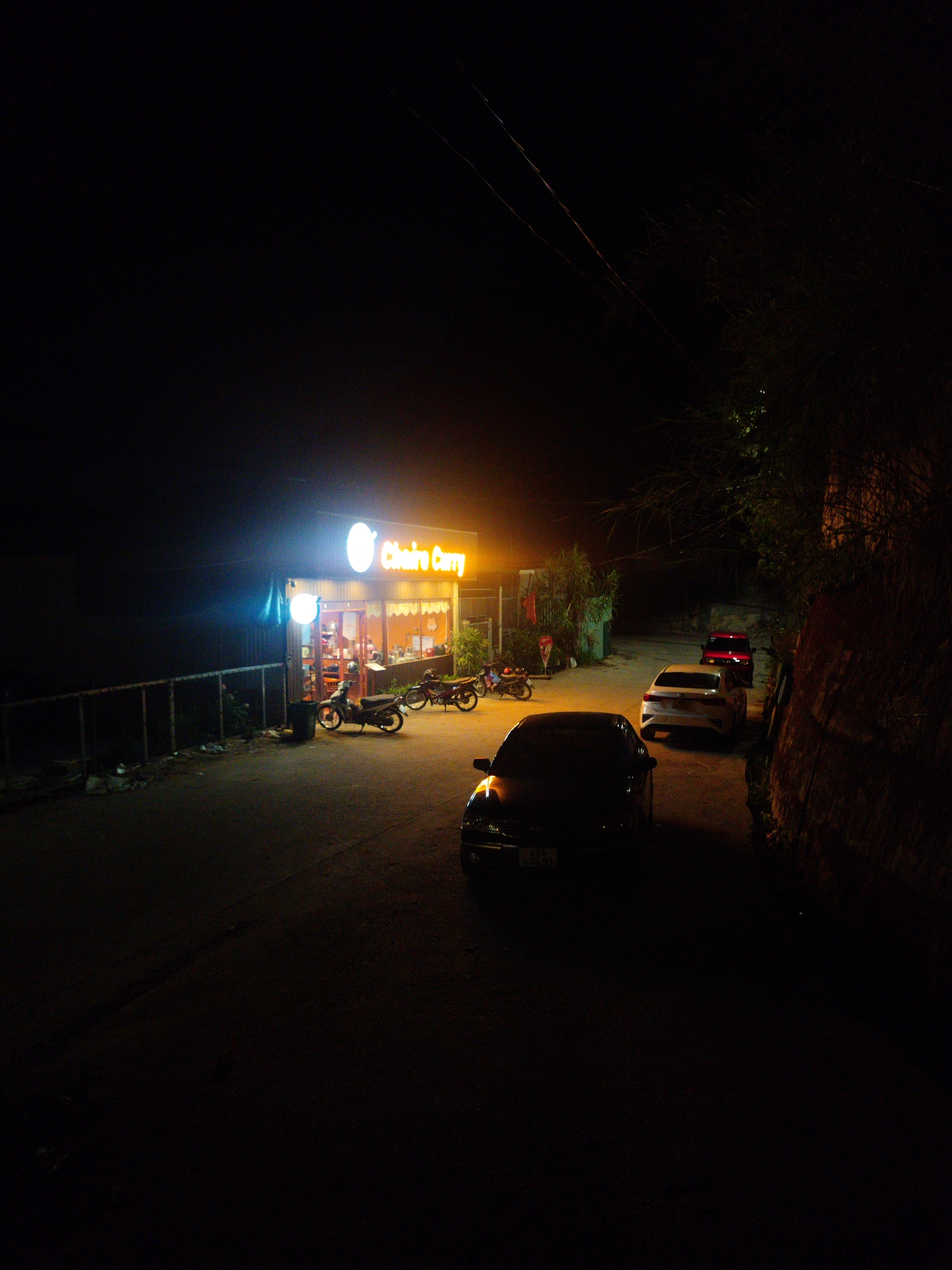 The image depicts a nighttime street scene in Da Lat, Vietnam. A brightly lit "Choiro Curry" restaurant is visible, with several parked motorbikes in front. Further down the street, there are three parked cars: a dark sedan in the foreground, a white sedan behind it, and a red car further in the distance. A metal fence runs along the left side of the street, while a dark, rocky embankment with vegetation is present on the right. Electrical lines are visible in the upper part of the image.