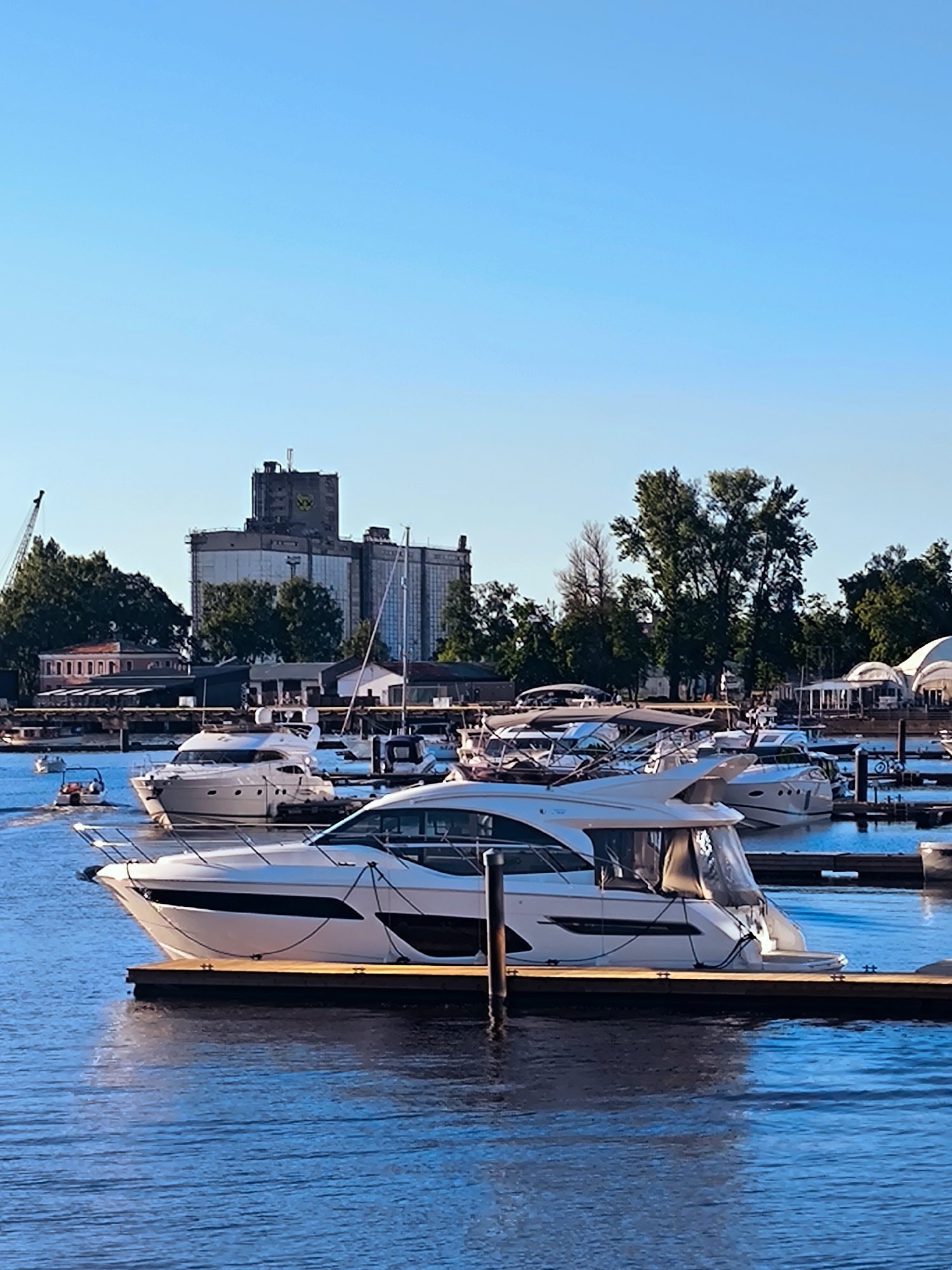 The image displays a marina on the Daugava River in Riga, Latvia, under a clear, bright blue sky, suggesting late afternoon or early evening. The river water is a deep blue with subtle surface ripples.
In the foreground and midground, multiple white luxury motor yachts are moored to wooden piers. The most prominent vessel in the center is a white motor yacht with dark windows and a tan-colored canvas enclosure over its aft cockpit, secured to a wooden dock. Another similar white motor yacht is partially visible behind it to the left. Further into the marina, additional white yachts are docked. On the far left, a small open motorboat, carrying at least one individual, is observed moving, producing a minor wake.
The background is dominated by the Rīgas Ostas Elevators (Riga Port Elevator), a large, multi-story industrial complex constructed of concrete. Its upper section is darker and features a distinct yellow circular logo. To the left of the elevator, a tall industrial crane is partially visible. Various smaller buildings, some with red roofs, are situated along the shoreline, interspersed with dense green deciduous trees. On the far right of the frame, several white, dome-shaped tent structures are discernible. No other individuals are distinctly visible or engaged in specific actions.