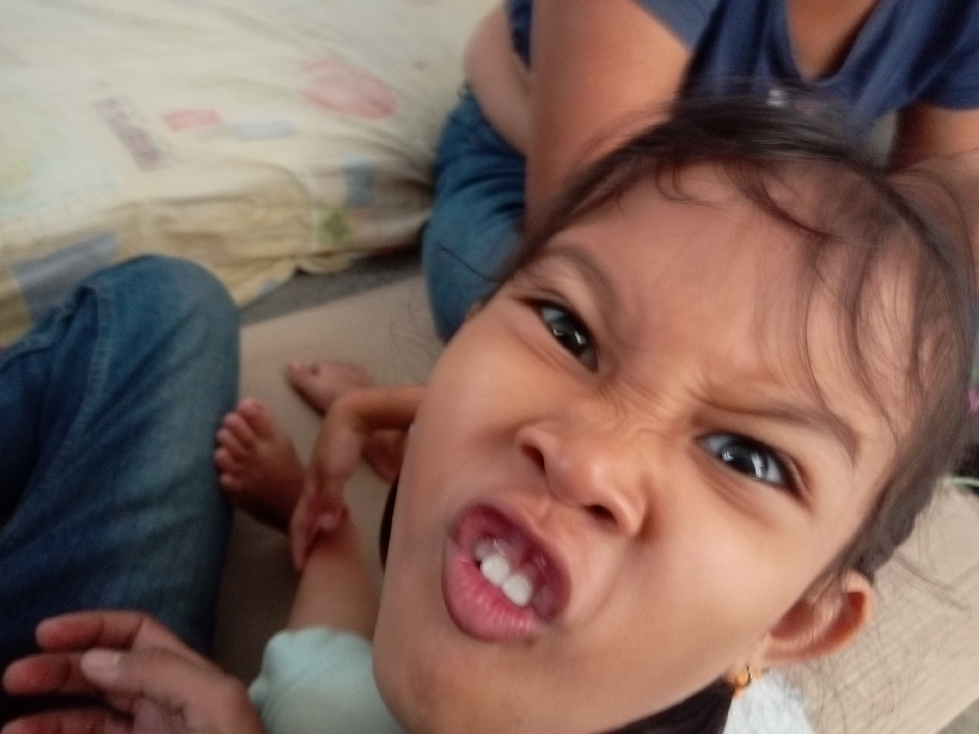 The image is a close-up, low-angle, candid photograph taken indoors, likely in a residential setting in Batam, Indonesia. The focus is on a young child, approximately 3-5 years old, with dark hair and brown eyes, making a funny face towards the camera. The child's mouth is open, revealing two front teeth, and their nose is scrunched. The child is wearing a light blue shirt.
To the left of the child, partially visible, is an adult's leg clad in blue jeans, suggesting the adult is seated or lying down. In the background, an adult's torso wearing a dark blue t-shirt is visible. The environment appears to be a room with a light-colored, possibly patterned, mattress or blanket spread out. The lighting is natural, indicating daylight. There are no overt indications of the specific location within Batam. The overall impression is of a casual, playful moment captured between family members.