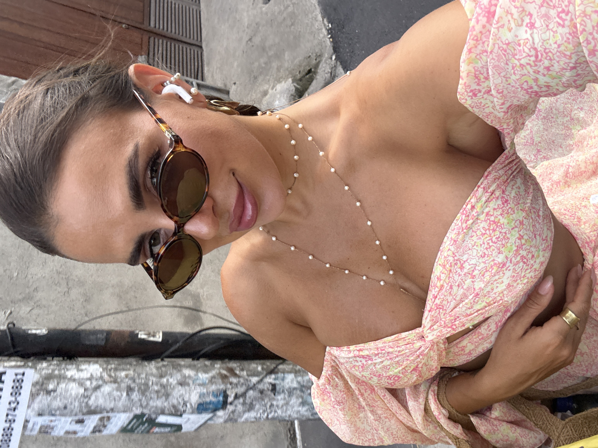 A woman wearing sunglasses and a pink floral patterned top is seen from a high angle. She is wearing a thin necklace with small pearl-like beads. An earbud and additional jewelry are visible on her left ear. The background shows a concrete surface with a visible telephone number and the urban environment. Based on the details provided, the location can be identified as North Kuta, Indonesia.