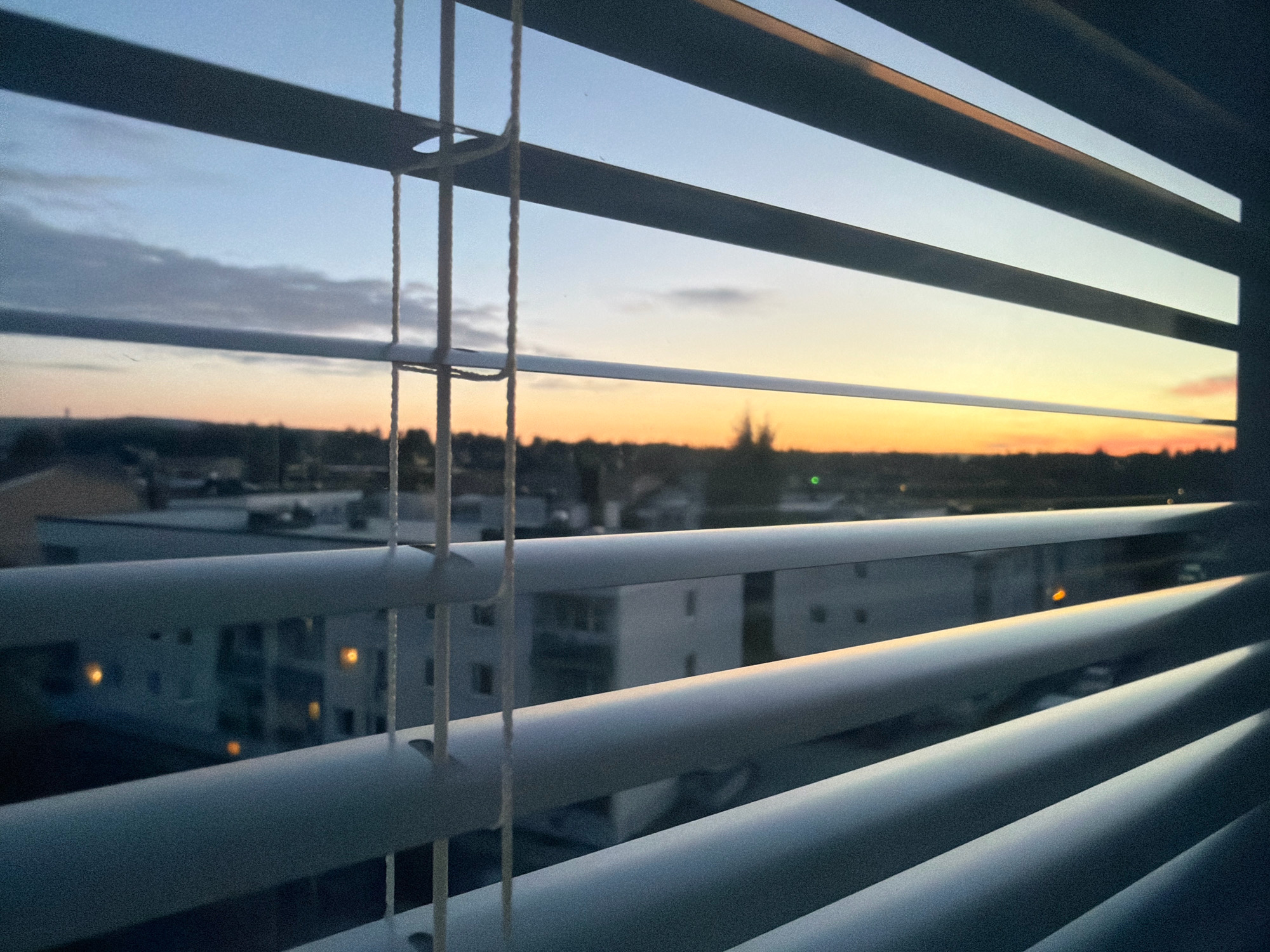 The image depicts a sunset viewed through horizontal window blinds. The blinds are white and partially obscure a cityscape. In the distance, the sky exhibits hues of blue, orange, and yellow, indicating the sun is low on the horizon. Buildings, including apartment complexes, are visible beneath the sky. The lights within some of the apartments are illuminated, casting a warm glow. The scene is captured from a vantage point likely within a residential building in Gällivare, Sweden, given the architectural style and the twilight illumination.