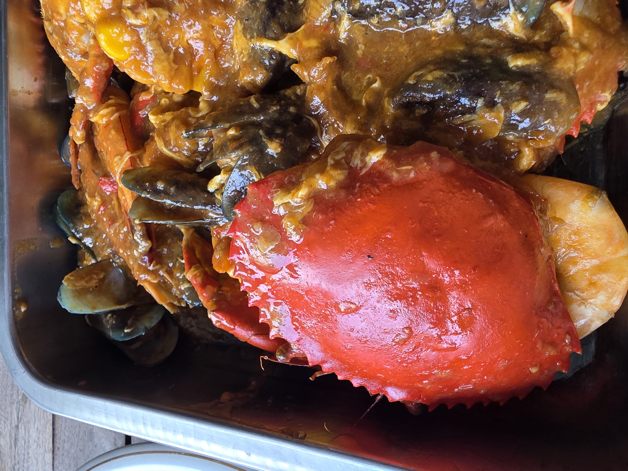A close-up, overhead shot shows a metal tray filled with a variety of seafood dishes, prominently featuring a large red crab shell in the foreground. The crab shell is glossy and covered in a thick, orange-colored sauce, possibly a chili or butter sauce, with specks of red chili visible. Other seafood, including mussels with their dark shells and streaks of blue, and what appear to be prawns or other shellfish, are also present and coated in the same sauce. The seafood is arranged within the confines of the rectangular metal tray, which rests on a wooden surface, likely a table. The lighting suggests an outdoor setting, possibly a restaurant in Seririt, Indonesia, where this seafood dish is being served.