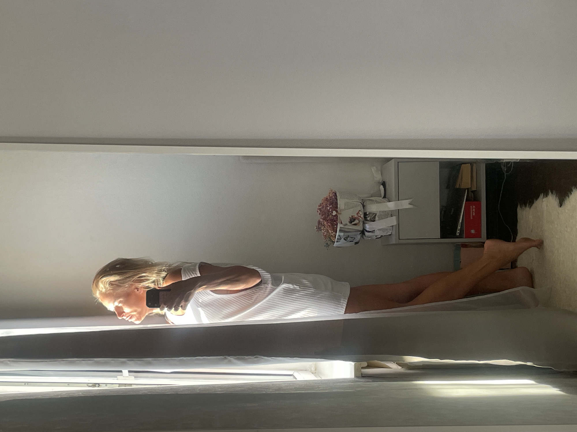 A woman stands in profile, facing a window, bathed in sunlight. She holds a phone in her right hand, angled as if taking a selfie. She wears a white, ribbed, off-the-shoulder dress that ends mid-thigh. Her hair is blonde and windswept. To her right, a small nightstand holds a basket of dried flowers. The scene appears to be indoors, possibly a bedroom, with a large mirror reflecting the woman and the room. The overall lighting suggests early morning or late afternoon sun. There is no indication of the location being in Baranovka, Russia.