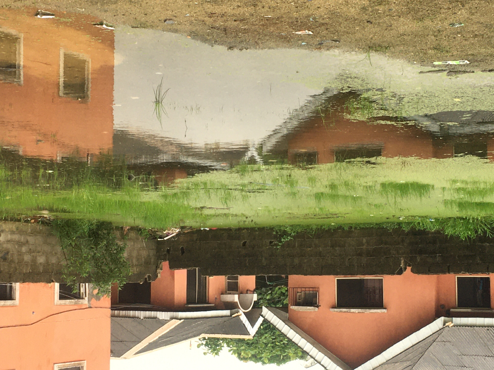 The image shows a reflection of a building in a puddle, inverted. The building is terracotta-colored with multiple windows and gray roofs. A concrete wall and greenery are visible between the building and the water's edge. The surrounding ground is dirt and debris-covered. The location is likely in Ado, Nigeria.