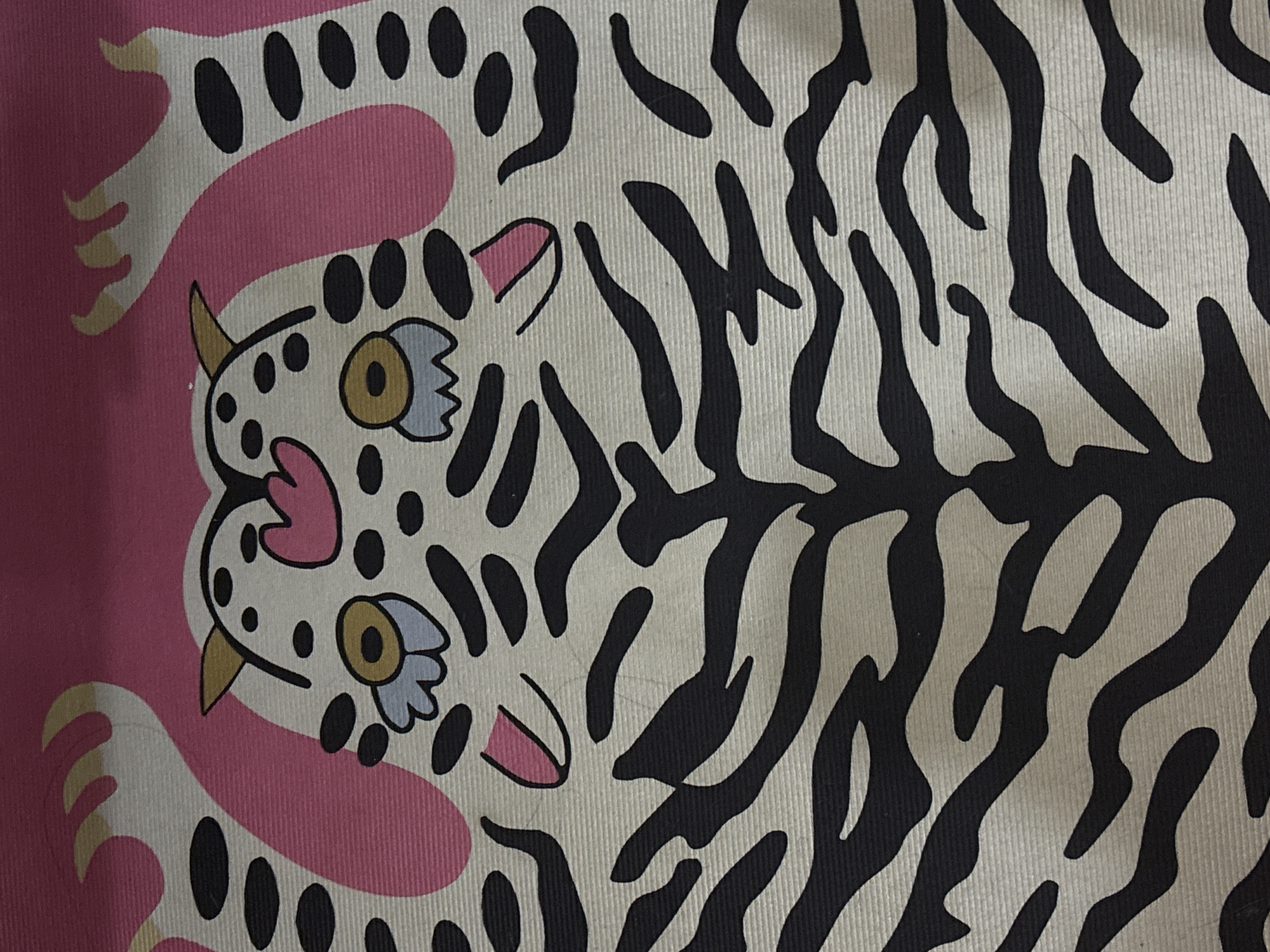 The image shows a close-up of a patterned fabric. The pattern includes a stylized depiction of two white tigers with black spots and stripes. The tigers have pink ears and noses, and yellow and blue eyes. One tiger is in a pink silhouette. The other tiger has black stripes. The fabric appears to have vertical ribs.