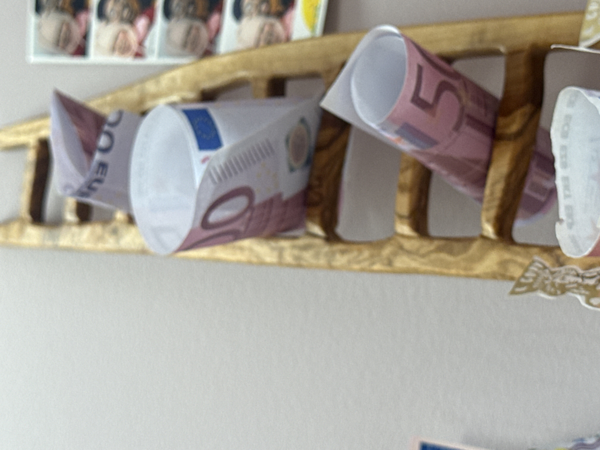 The image shows a close-up of a wooden ladder with several rolled-up euro banknotes placed on its rungs. The banknotes are primarily pink and feature numbers like "50" and "100" prominently displayed. Some of the banknotes also show a blue European Union star symbol. To the right of the ladder, a framed collage of photographs is visible, displaying multiple candid shots of people, some with pink hues and others in color. The overall setting appears to be indoors, with a plain, light-colored wall as the background. The exact location in Sankt Andrä-Wördern, Austria, is not determinable from the provided image.