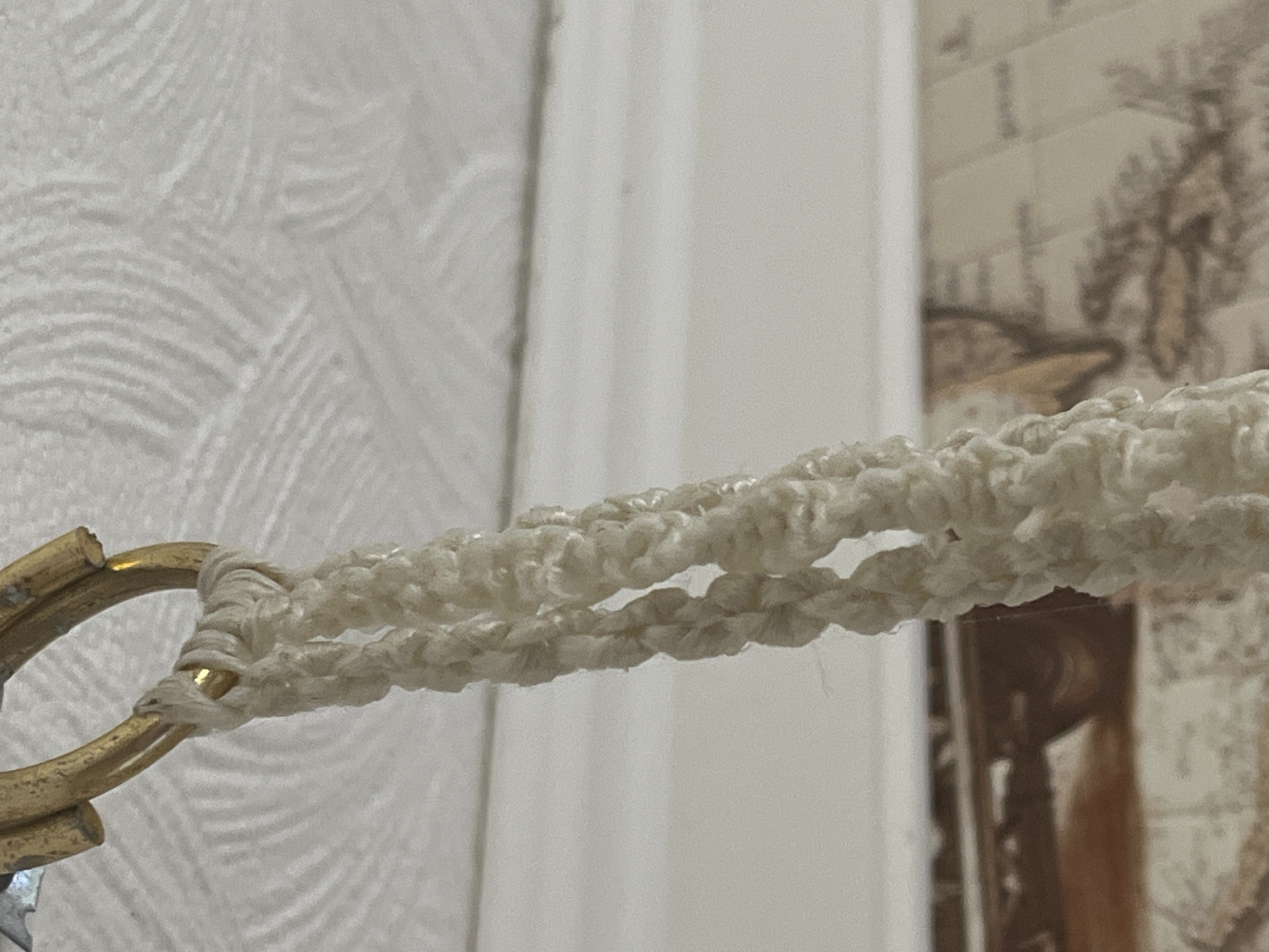 This is a close-up, low-angle, indoor shot of a macrame plant hanger made of thick, cream-colored yarn, attached to a golden ring that is screwed into a textured, white ceiling. The macrame forms a braided pattern with several openings. The background is blurred, but appears to show a wall with a map or patterned wallpaper and possibly a shelf or table with some objects. There are no people or animals visible. The scene is static and still. The location is not identifiable from the image itself, and therefore cannot be specified as Krasnyye Barrikady, Russia.