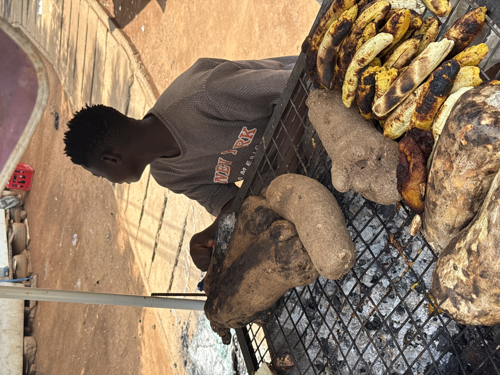 A street food vendor is roasting plantains and yams on a charcoal grill in Abuja, Nigeria. The vendor is a young, dark-skinned man wearing a gray shirt with a "New York" print. He is positioned to the left of the grill, tending to the food. On the grill, charred plantains are piled up towards the upper right, with yams of varying sizes scattered throughout. The grill's metal grate is visible, showing charcoal remnants underneath. The background includes a sunlit sidewalk and a glimpse of a red object and other equipment.