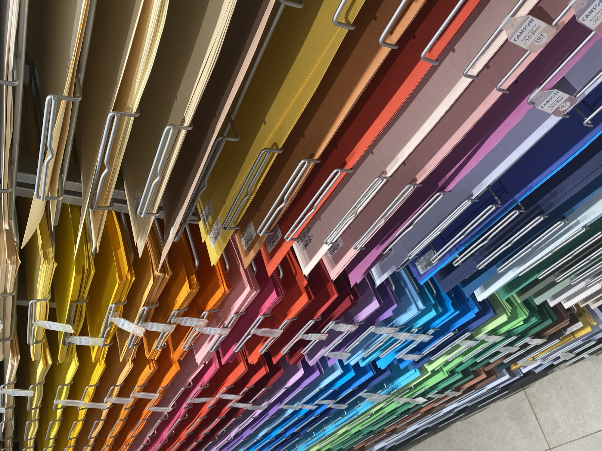 The image shows a display of colorful paper sheets organized by color and stacked on metal shelving units. The paper ranges from pale yellow and orange to vibrant reds, pinks, purples, blues, and greens. Some sheets have tags attached to the shelving, with text that appears to be branding and product information. The shelving units are arranged in rows, suggesting a retail environment. The lighting suggests an indoor setting, likely a store or art supply shop. The floor in the lower left corner appears to be tiled. No people are visible in the image. The exact location in Warsaw, Poland is not identifiable from the visual information provided.