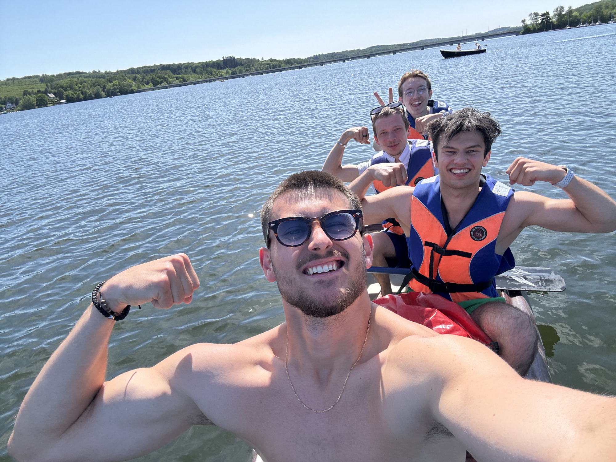 Four people pose on a boat. The man at the front, shirtless and wearing sunglasses and a gold chain, flexes his bicep. Behind him, three other men are visible, each wearing a life jacket. The man immediately behind the first man also flexes. The second man makes a "peace" sign. The fourth man is partially obscured. The background reveals a body of water, presumably the Möhnesee. In the distance, forested areas and a bridge are visible. A small boat with two people is also visible in the water.