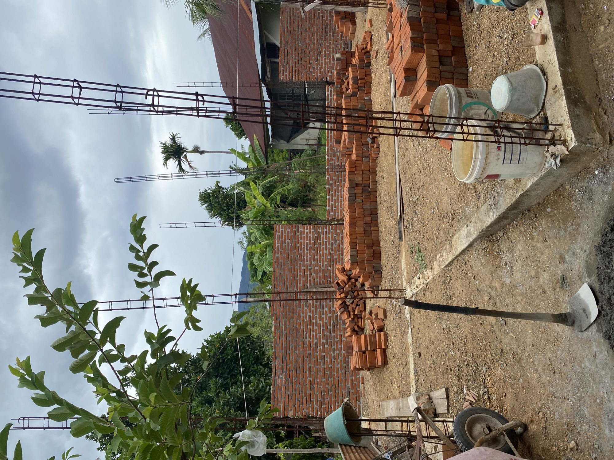 An outdoor, ground-level, medium shot captures an under-construction building. In the foreground, a shovel with a grey handle and a metal head is stuck vertically into the dirt. To the left, the wheel of a pink wheelbarrow is visible. In the middle ground, numerous stacks of red bricks are arranged on the dusty ground, some forming a partial wall. Several vertical metal rebar columns are also present, some extending high into the sky. In the background, a mature green leafy tree is on the left, and a tropical palm tree is to the right of the structure. A simple house with a corrugated metal roof is situated behind the construction site. The sky is overcast with gray clouds.