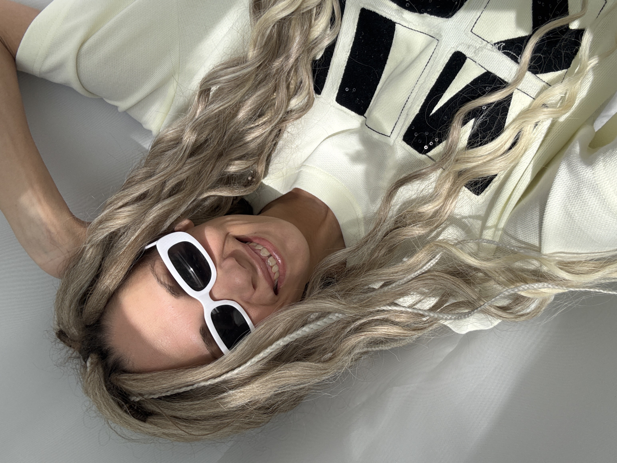 A person lies on their back in what appears to be an indoor setting, dressed in a light yellow top with black and white detailing. They are wearing white-framed sunglasses and smiling, with long, wavy blonde hair spread around their head. No specific details identify the location as Rusanovo, Russia. The background is a light, solid color.