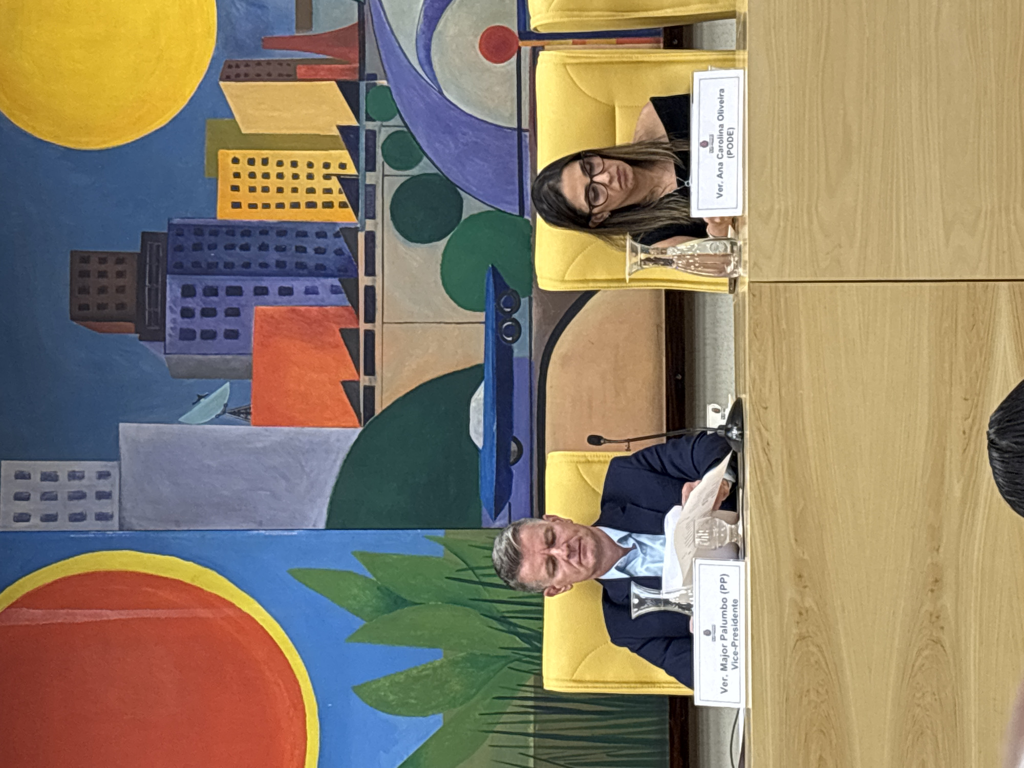 The image shows two people, a man and a woman, seated at a table with nameplates identifying them as Ver. Major Palumbo (PP) and Ver. Ana Carolina Oliveira (PODE). They are likely at a meeting or conference, as indicated by their formal attire and the presence of microphones. Behind them is a large, colorful mural depicting a stylized cityscape, possibly representing the city of São Paulo, Brazil. The mural includes geometric shapes and bold colors, with buildings, a sun, and abstract forms.