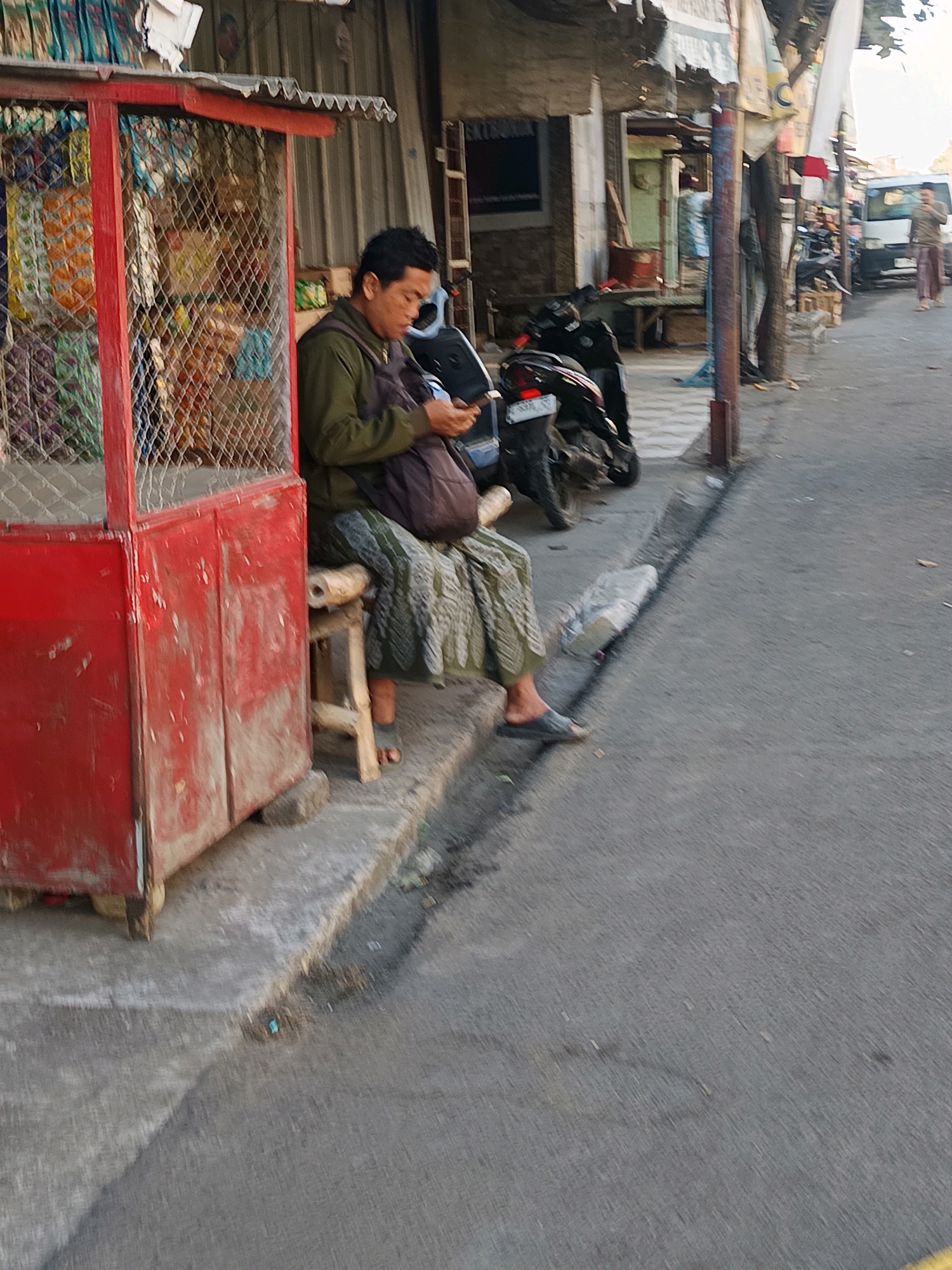 A man is sitting on a small stool in front of a red roadside stall. He is wearing a green jacket and a patterned cloth skirt. He is looking down at his phone. Behind him, there is a motorcycle. The stall is filled with various items, including bags of snacks. The scene appears to be a street in Blega, Indonesia, with a paved road and buildings in the background.