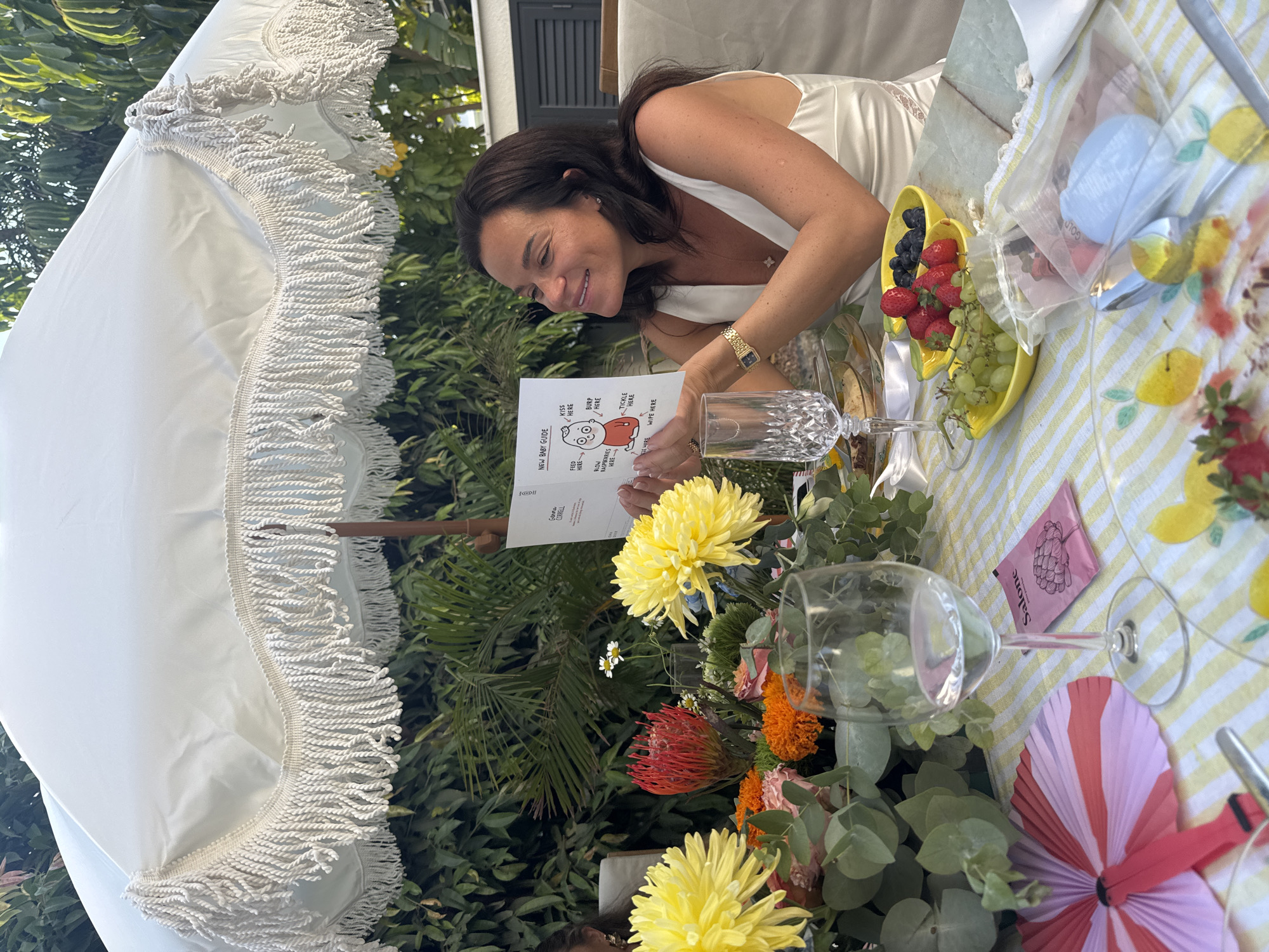 A woman is seated at a table outdoors, under a fringed white umbrella, holding a "New Baby Guide" card. She is smiling and looking at the card, which depicts a cartoon baby with labels for actions such as "Kiss Here," "Burp Here," and "Tickle Here." The table is set with a yellow and white striped tablecloth, a floral arrangement, glasses of water and wine, bowls of fruit (strawberries, blueberries, and grapes), and a decorative fan. The woman is wearing a white satin dress and a gold watch. In the background, lush green foliage suggests a garden setting. The scene appears to be a baby shower or a celebratory gathering. The location is described as Agios Tychona Community, Cyprus.