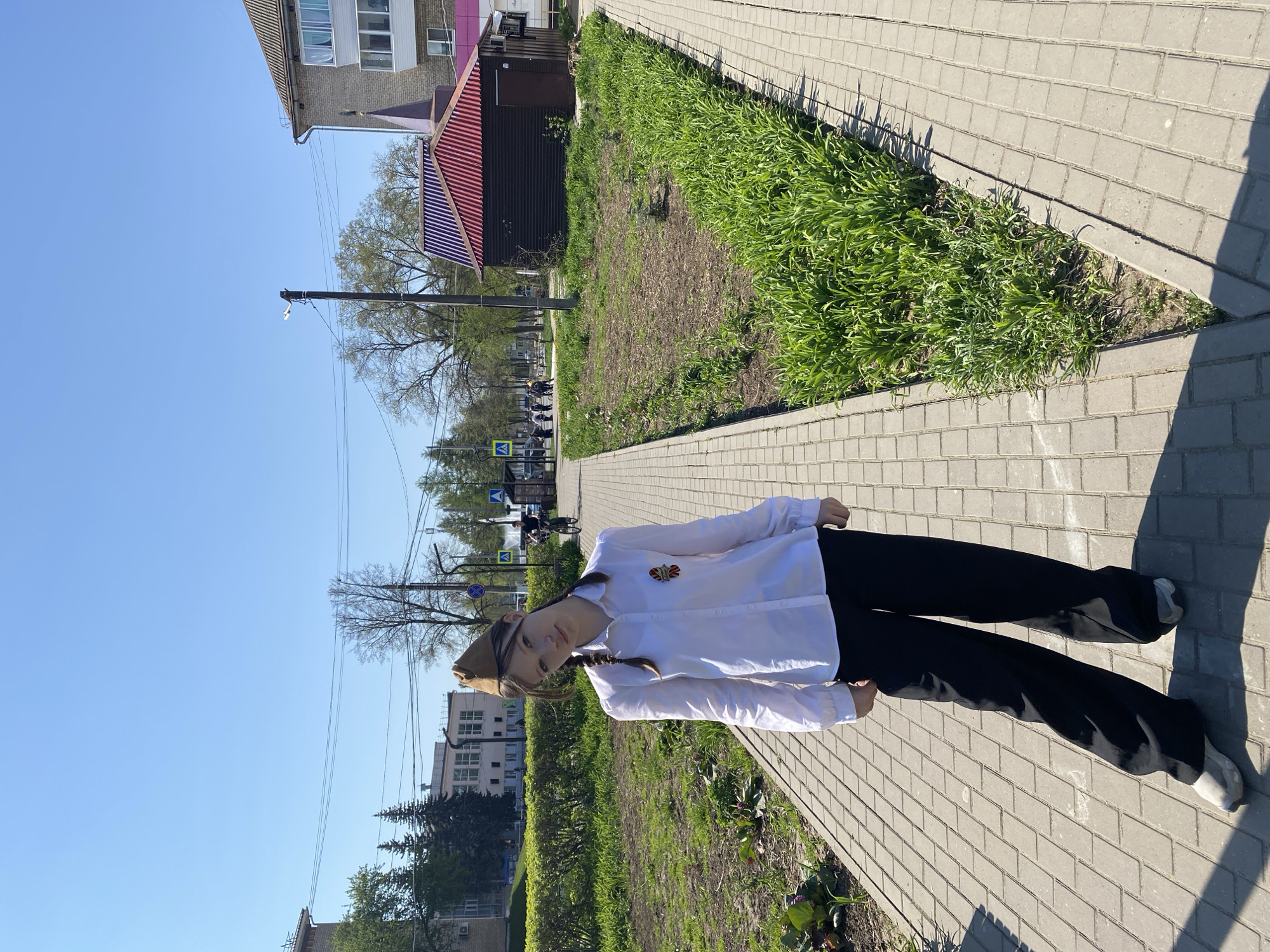 A young woman stands on a paved walkway in what appears to be Chaikovsky, Russia, on a sunny day. She is wearing a white button-down shirt, black pants, and a military-style cap. Her hair is braided and she is wearing a pin on her shirt. In the background, there are buildings, trees, and a street with cars. The walkway is made of rectangular grey pavers, and there is grass growing along the side.
