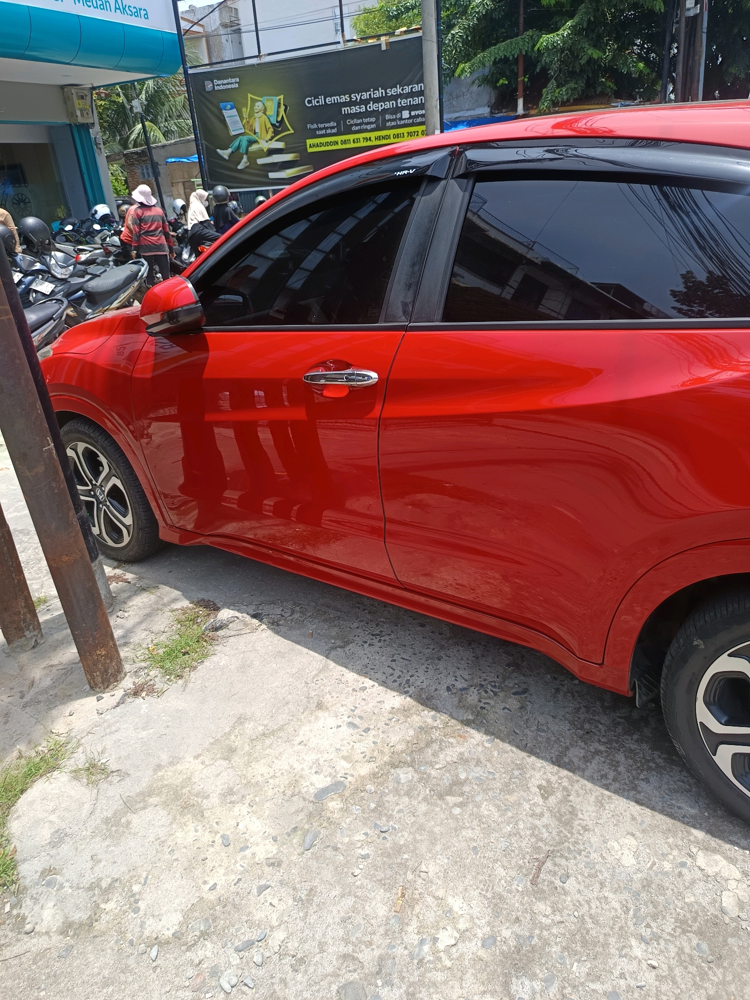 The image shows a red car parked on a concrete surface in front of a building. There are several motorcycles parked behind the car, and a few people are visible near the motorcycles. The building appears to be a commercial establishment, possibly a shop or an office. A banner is displayed on the building with text in Indonesian. The overall environment suggests an urban setting in Percut Sei Tuan, Indonesia. The lighting indicates it is daytime, with some shadows cast by the car and other objects.