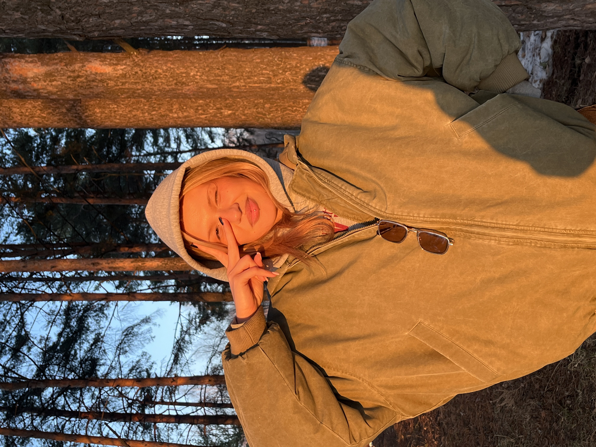 A blonde woman in a gray hooded sweatshirt and a green jacket poses with a peace sign in front of her face. Sunglasses are tucked into the jacket. The background shows a forest with a visible blue sky through the trees. The location is likely Chaikovsky, Russia.