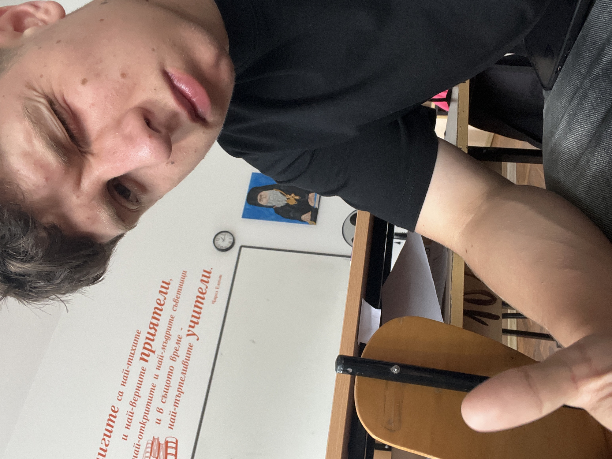A young person, likely male, takes a selfie in what appears to be a classroom in Zlatograd, Bulgaria. The individual, dressed in a black short-sleeved t-shirt, is positioned in the foreground, looking directly into the camera with one eye squinted or winking and lips slightly parted in a casual expression. Their right arm, with a visible vein, rests on a wooden desk. A hand, possibly their left, is seen holding or resting near a wooden chair seat in the bottom right corner.
The setting includes a white board on the wall in the background, above which a small analog clock with a black rim is mounted. To the left of the whiteboard, a small framed picture or icon of a robed figure, possibly a saint or monk with a golden halo, is displayed. A poster or decal with Cyrillic text is prominently featured on the wall to the left of the whiteboard.
The visible text, confirmed by OCR, reads: "Книгите са най-тихите и най-верните приятели, най-откритите и най-мъдрите съветници и в същото време най-търпеливите учители. Чарлз Елиът" (Books are the quietest and most faithful friends, the most open and wisest counselors and at the same time the most patient teachers. Charles Eliot). This quote reinforces the educational environment. The image is taken indoors, with no specific time of day or weather discernible.