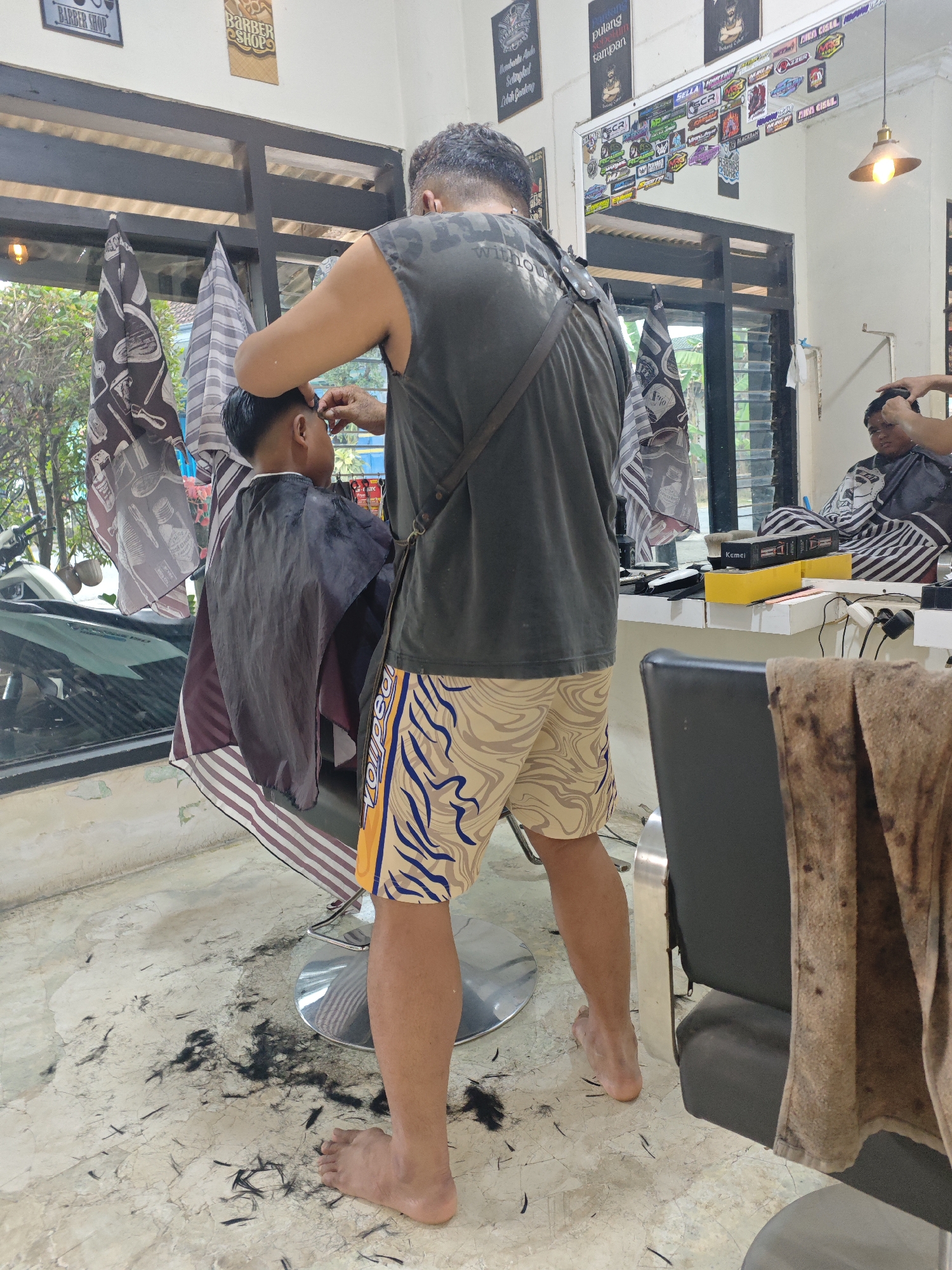 This image captures a barber giving a haircut to a young boy in a barber shop. The barber, a man with short, dark hair, is wearing a sleeveless dark shirt and patterned shorts. He is standing behind the boy and using scissors to trim the boy's hair. The boy is seated in a barber chair, wearing a black cape. The barber shop appears to be in Sarongan, Indonesia, as indicated by the overall aesthetic and signage visible in the background, which features Indonesian text and popular local decals. The floor is littered with cut hair, and a motorcycle is partially visible through the open doorway to the left, suggesting an outdoor or semi-outdoor setting. In the background, a mirror reflects another customer in a barber chair and various barber shop tools and equipment, including clippers and product boxes. The lighting suggests it is daytime, with natural light coming from the open doorway and an overhead lamp.