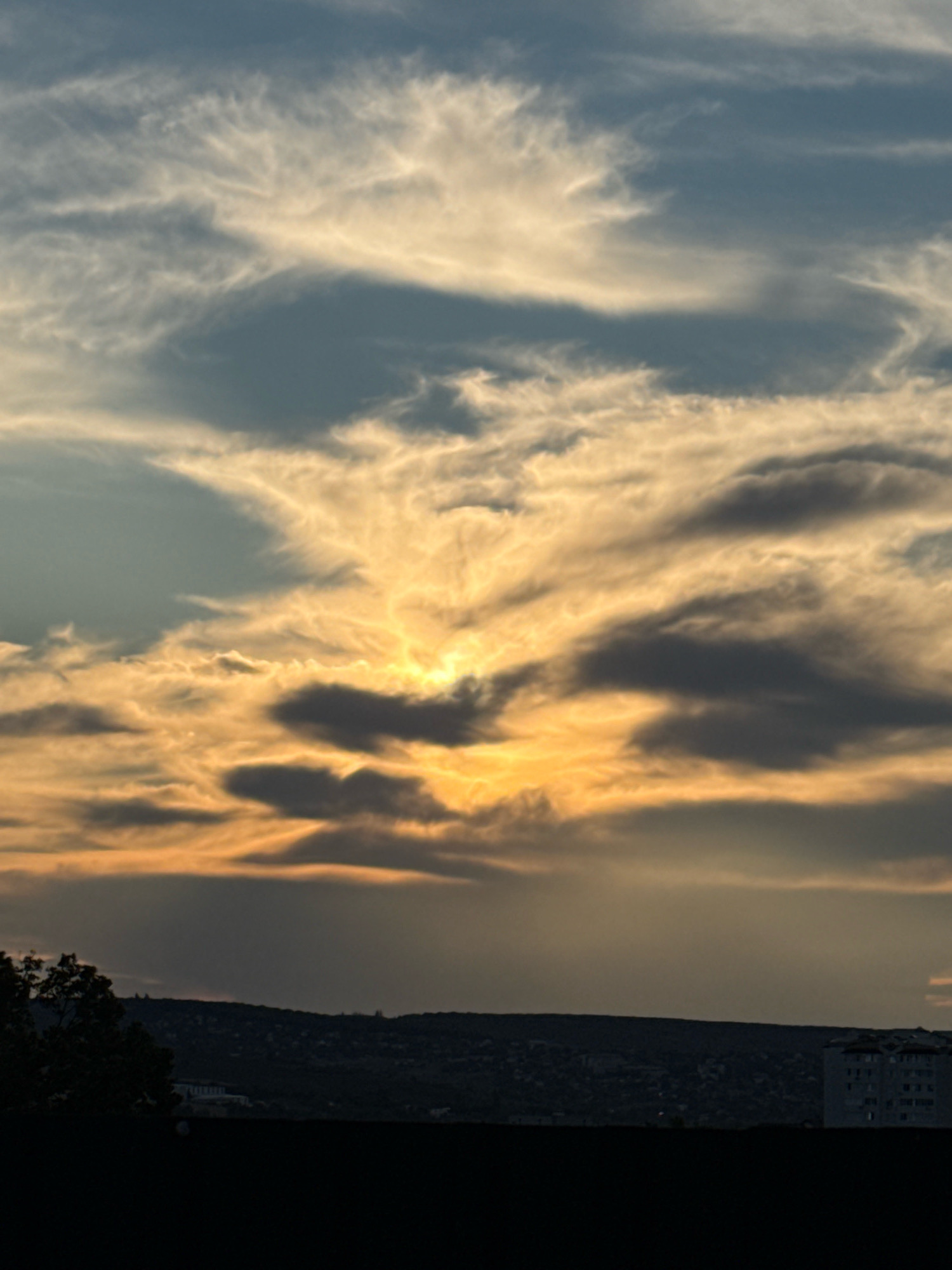 The image captures a dramatic sunset over the city of Ialoveni, Moldova. The sky is filled with wispy, striated clouds illuminated by the setting sun, which is partially obscured by darker, more dense cloud formations. The colors range from a pale blue at the top of the frame to warm oranges and yellows closer to the horizon. Below the sky, a dark silhouette of a hilly landscape is visible, dotted with what appear to be buildings and trees, suggesting a semi-urban or suburban setting. The foreground is dominated by a very dark, blurry area, possibly a rooftop or a similar elevated surface. On the right side of the landscape silhouette, a multi-story building with visible windows stands out against the darkening terrain, its architectural details somewhat obscured by the low light. The overall impression is of a serene but visually striking end to the day.