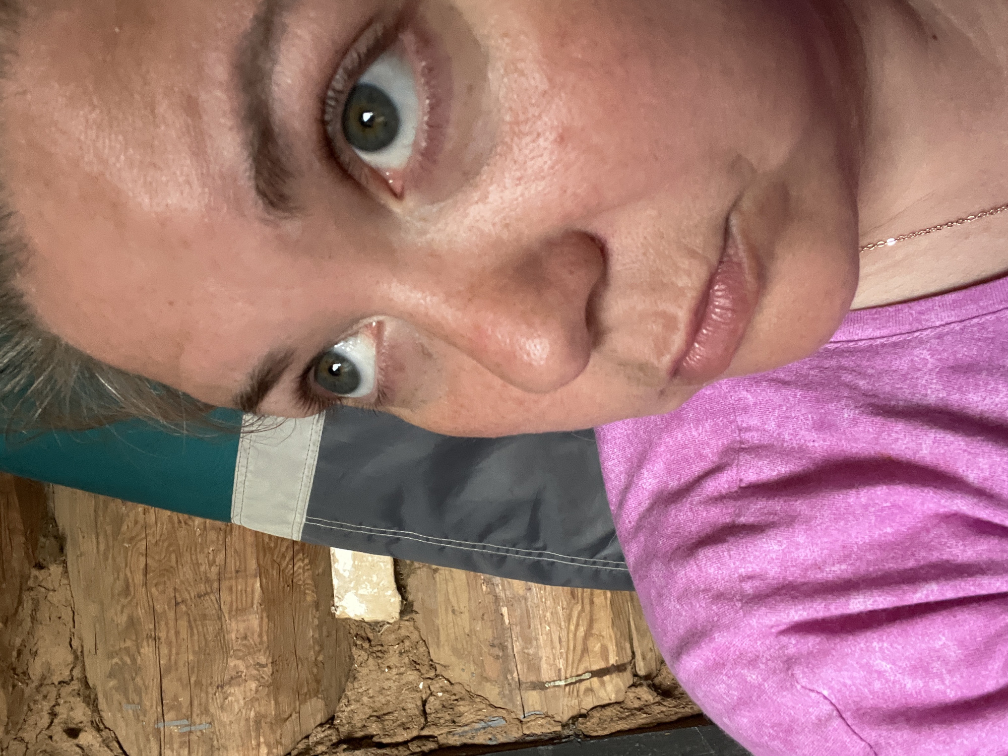 An extreme close-up, rotated selfie features a person with light skin, green/hazel eyes, and light hair, looking down and to the left. The upper part of their face, including the forehead, eyes, nose, and lips, is visible. They wear a pink, textured top and a delicate gold-toned chain necklace. The background shows rustic, rough-hewn wooden planks or logs, along with a piece of fabric that appears to be a flag or banner, displaying teal, white, and dark gray sections. Some dirt or debris is also present on the wooden surface. The lighting suggests an indoor setting. The person's expression is neutral, and no specific activity other than taking the selfie is evident. There is no visible text in the image. The location context provided is Фоки, Russia.
