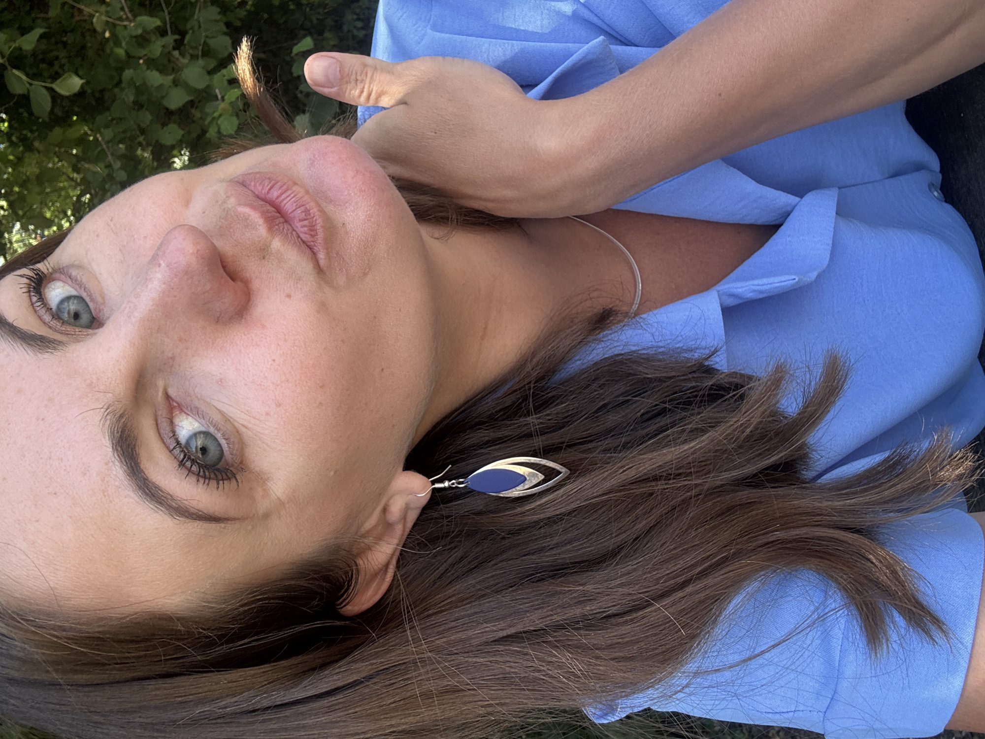 A close-up, waist-up, medium shot of a woman from the chest up is featured in this photograph. The woman is looking at the camera with her left eye visible, and her lips pursed as if about to kiss. She has brown hair, fair skin, and blue eyes. She is wearing a blue collared shirt and a silver necklace. A silver earring is visible in her left ear. The background consists of greenery and trees, suggesting an outdoor setting. The image is taken during the day, with natural light illuminating the subject. There is no specific indication of the location within Coteaux du Lizon, France.
