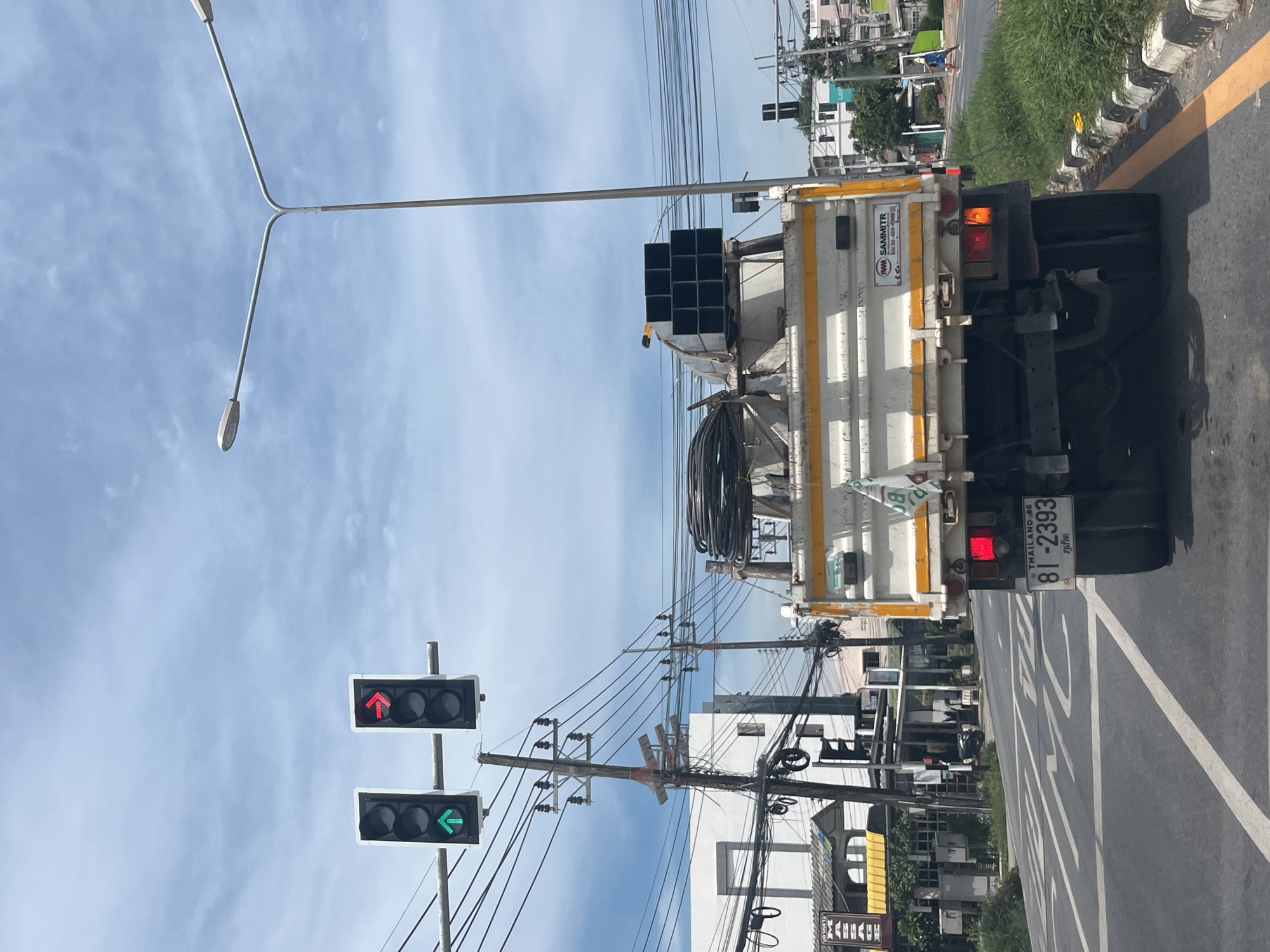 In Rawai, Thailand, on a bright, partly cloudy day, a white dump truck with a yellow stripe and metal beams is positioned in the foreground, facing away from the viewer. The truck's license plate reads "81-2393." To the left of the truck, a traffic light displays a green upward arrow, and a second traffic light shows a red right arrow. Overhead, power lines and streetlights are visible against the sky. In the background, buildings, trees, and a pedestrian are present.