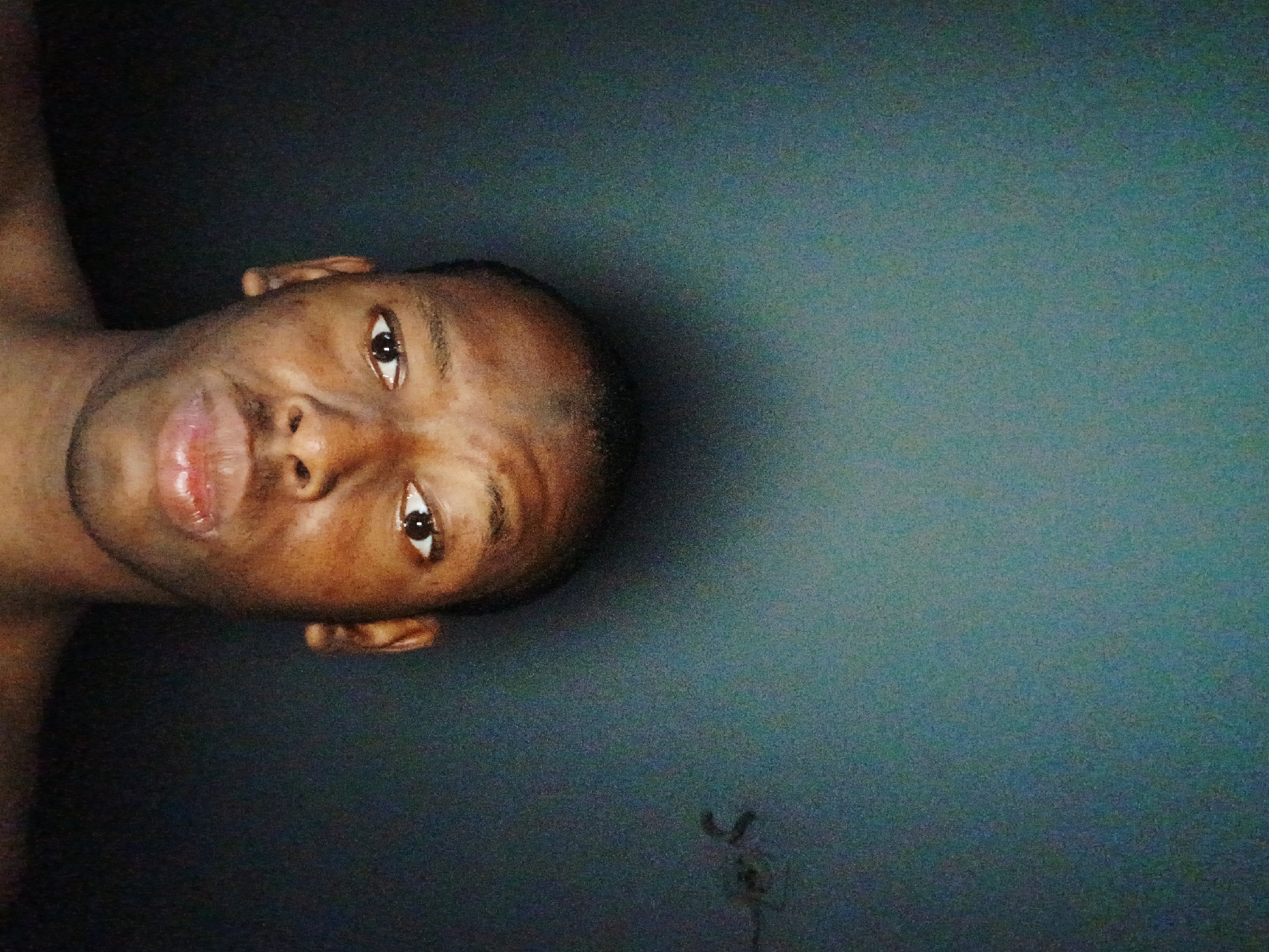 A close-up photograph shows a Black male with short-cropped hair, facing the camera, and positioned on the left side of the frame against a textured, dark blue-gray background. The background is featureless, except for an indistinct shadowy patch near the bottom right. The subject has dark eyes, slightly parted lips, and appears shirtless with a portion of his shoulder visible. The photograph appears to be taken in a dimly lit environment. The image's orientation is rotated 90 degrees counter-clockwise. The scene is unidentifiable in terms of geographic location.