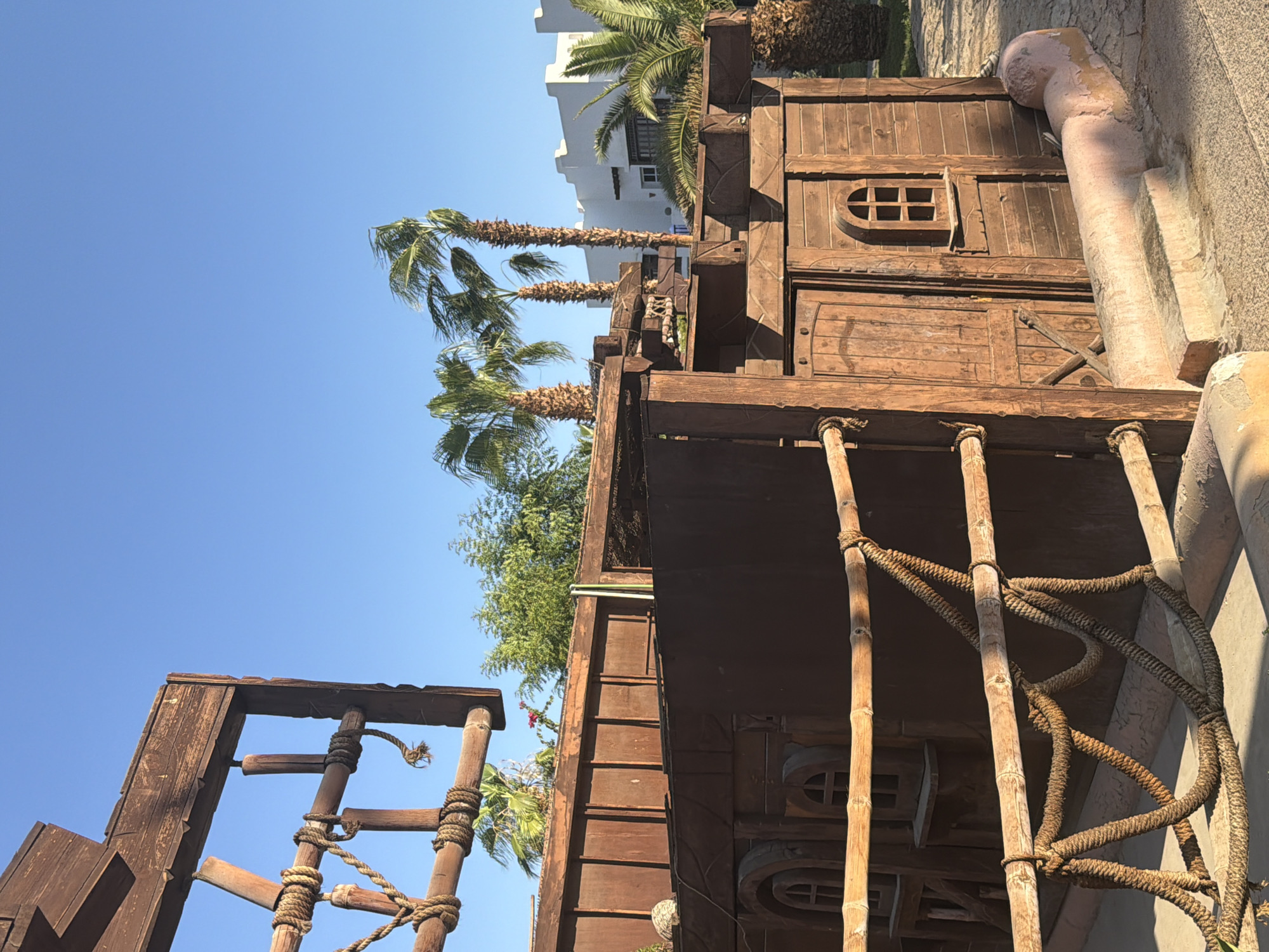 The image shows a wooden structure designed to resemble a pirate ship. It is situated outdoors in a sunny environment with palm trees and modern white buildings in the background, indicative of a resort or hotel in Sharm El Sheikh, Egypt. The wooden ship structure features planking, a door, and windows with arched tops. A rope railing with bamboo supports is visible in the foreground, adding to the nautical theme. The ground appears to be paved. The sky is clear and blue.