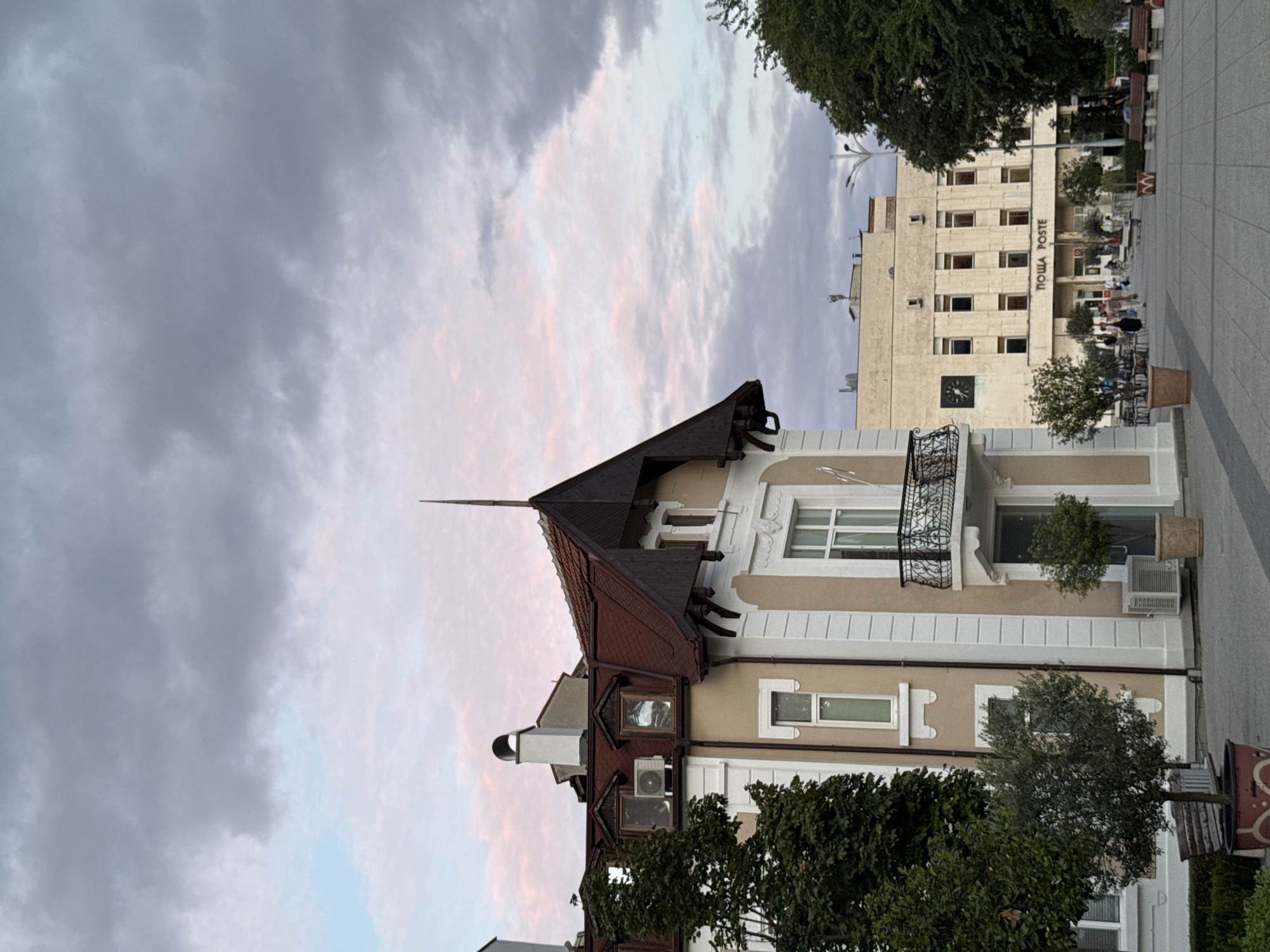 This is a photograph taken from a pedestrian plaza in the city of Plovdiv, Bulgaria. In the foreground, a beige and white building with a red tiled roof and a balcony with ornate metal railing is visible on the left. Trees are planted in pots around the building. Further back and to the right, a larger, light-colored, multi-story building with a prominent sign that reads "ПОЩА POSTE" is present. This building appears to be a post office. People are walking and sitting on benches in the plaza. The sky above is cloudy with some pink hues suggesting either sunrise or sunset. The overall scene depicts an urban environment with civic buildings and public space.